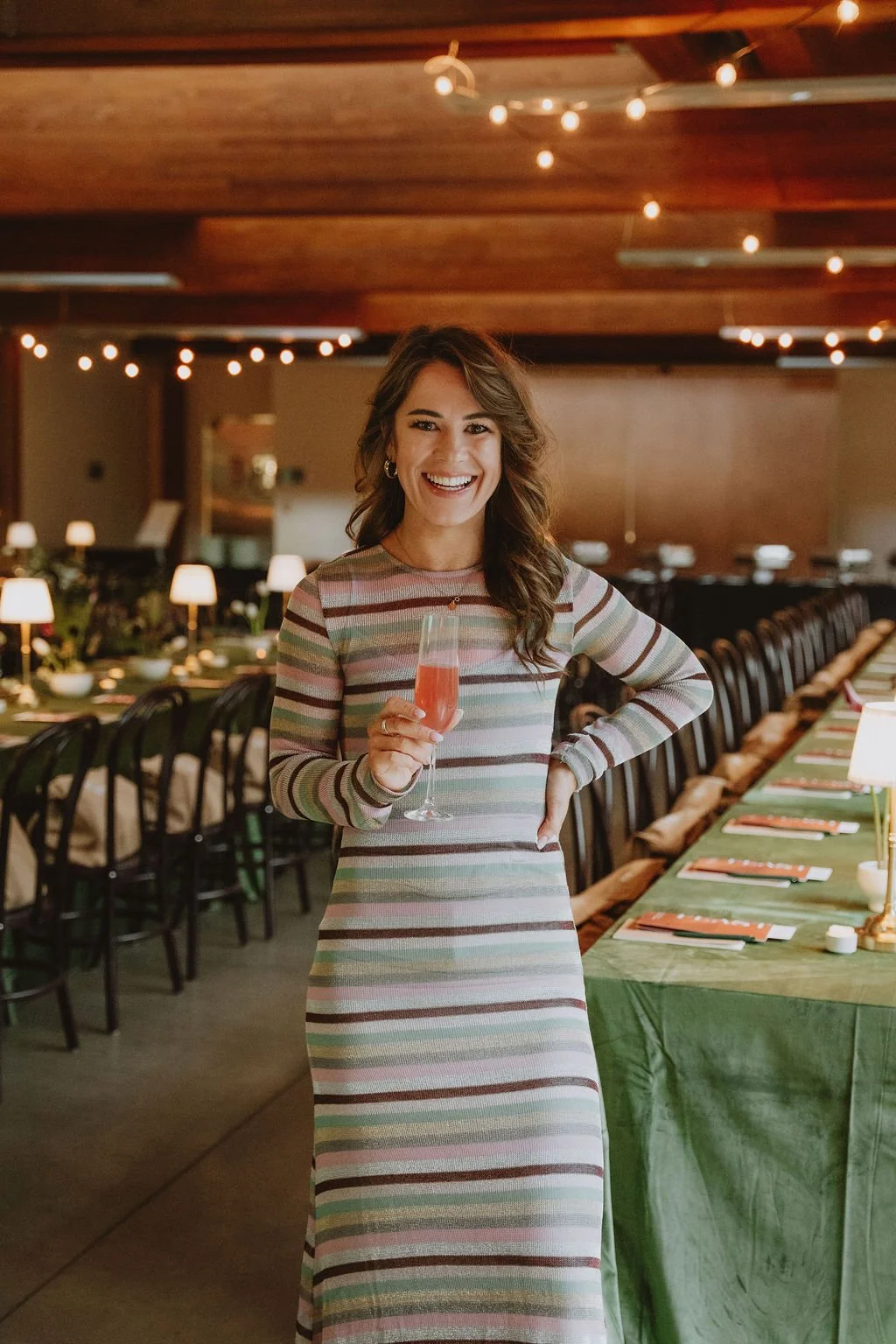 A woman in a striped dress smiling and holding a glass of pink drink in a decorated indoor celebration venue with string lights and table settings.