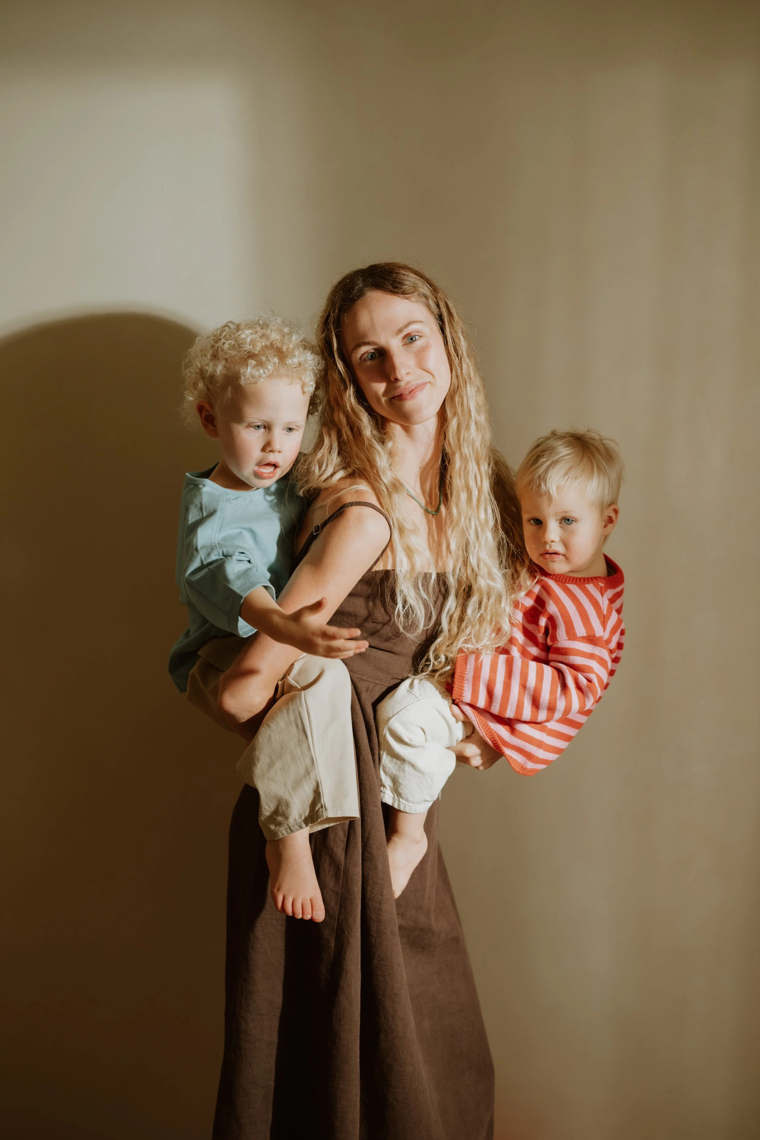 A young woman with long, curly blonde hair is holding two small children, one on each side. The child on her left has curly blonde hair and is wearing a light blue shirt, while the child on her right has straight blonde hair and is dressed in a red a