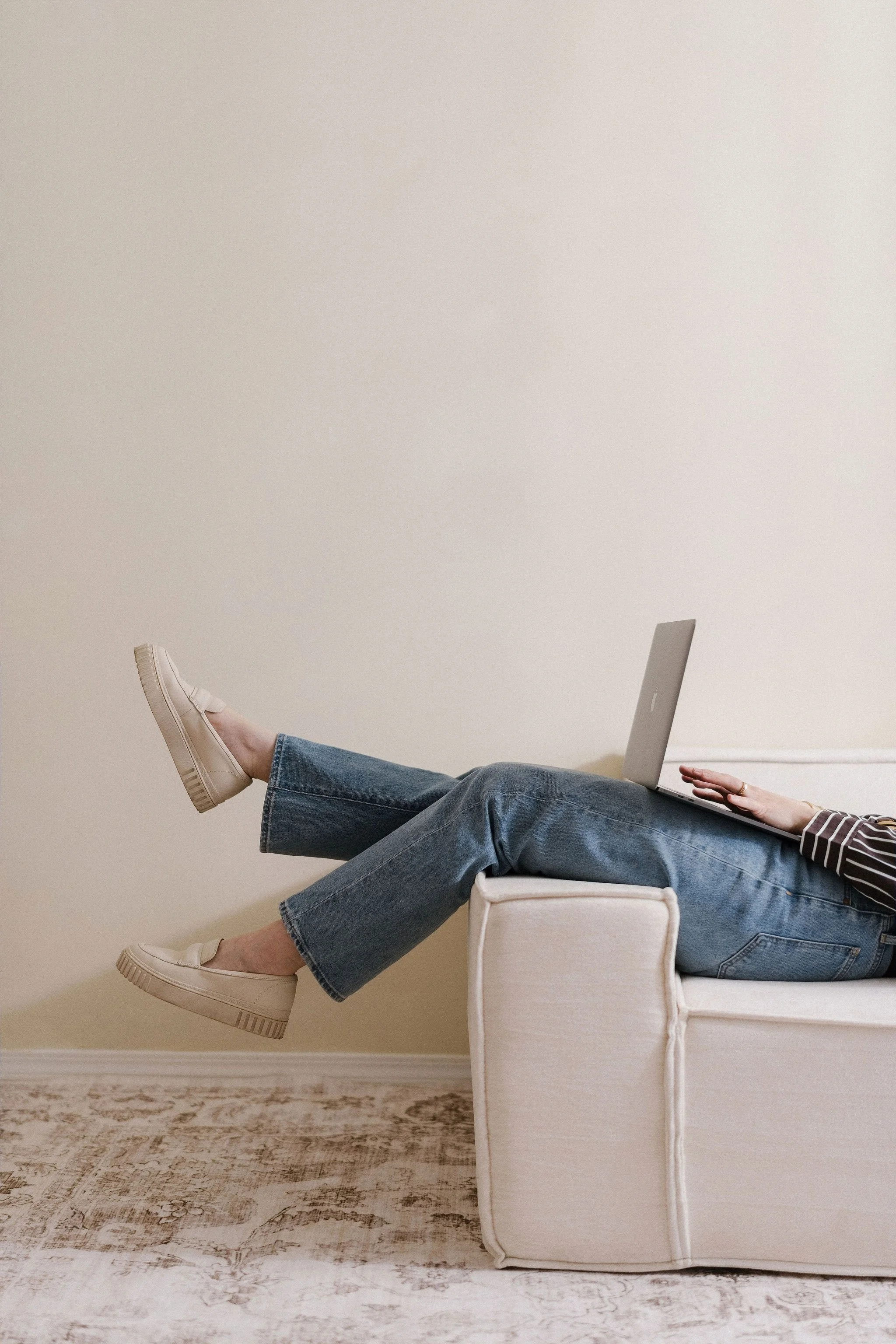 Person lying on a white armchair with legs extended, wearing beige sneakers, jeans, and a striped shirt, using a laptop, against a plain light-colored wall and a patterned rug.