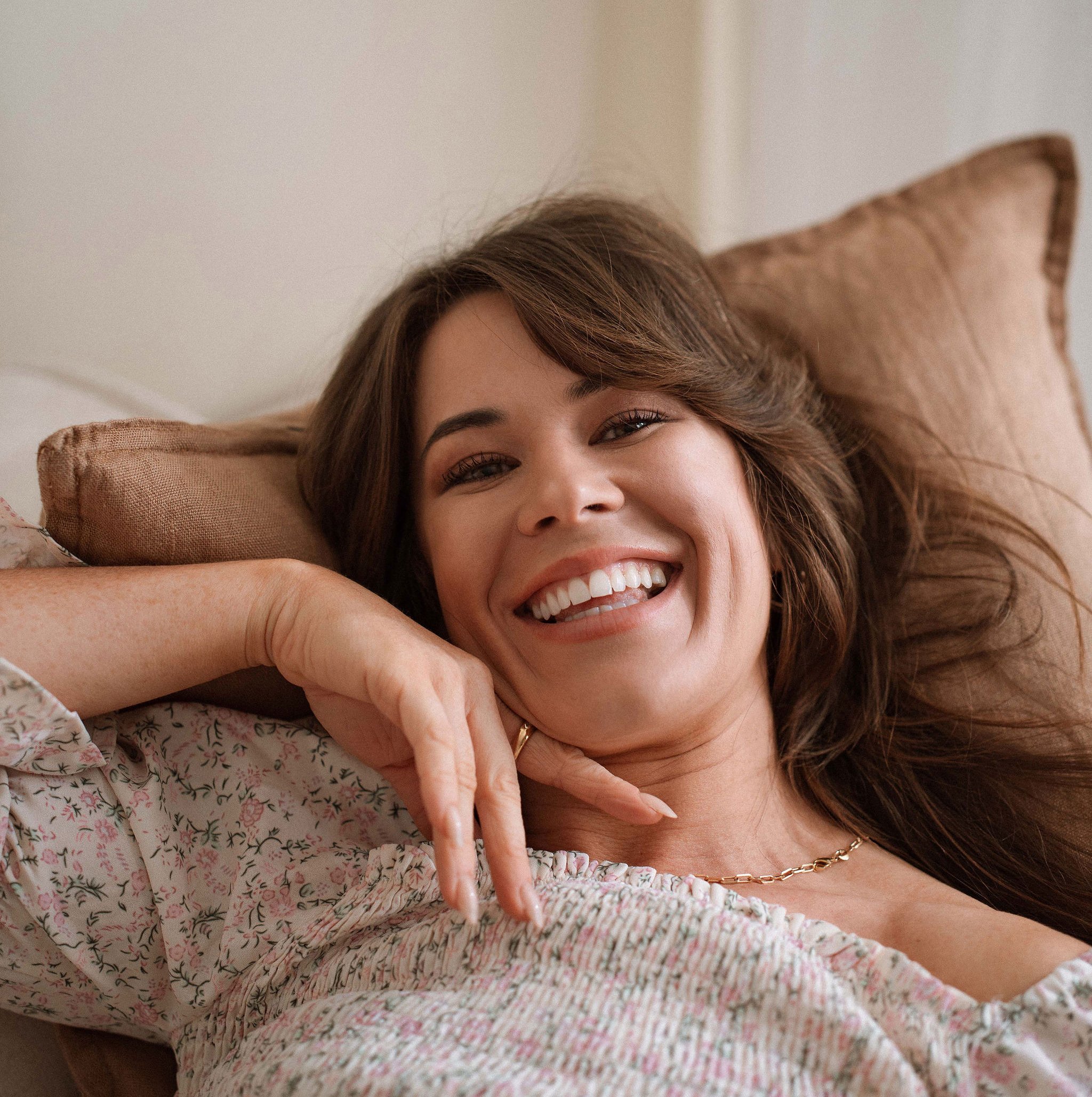 A woman with long brown hair smiling, lying on a bed with light-colored bedding and a tiled headboard.