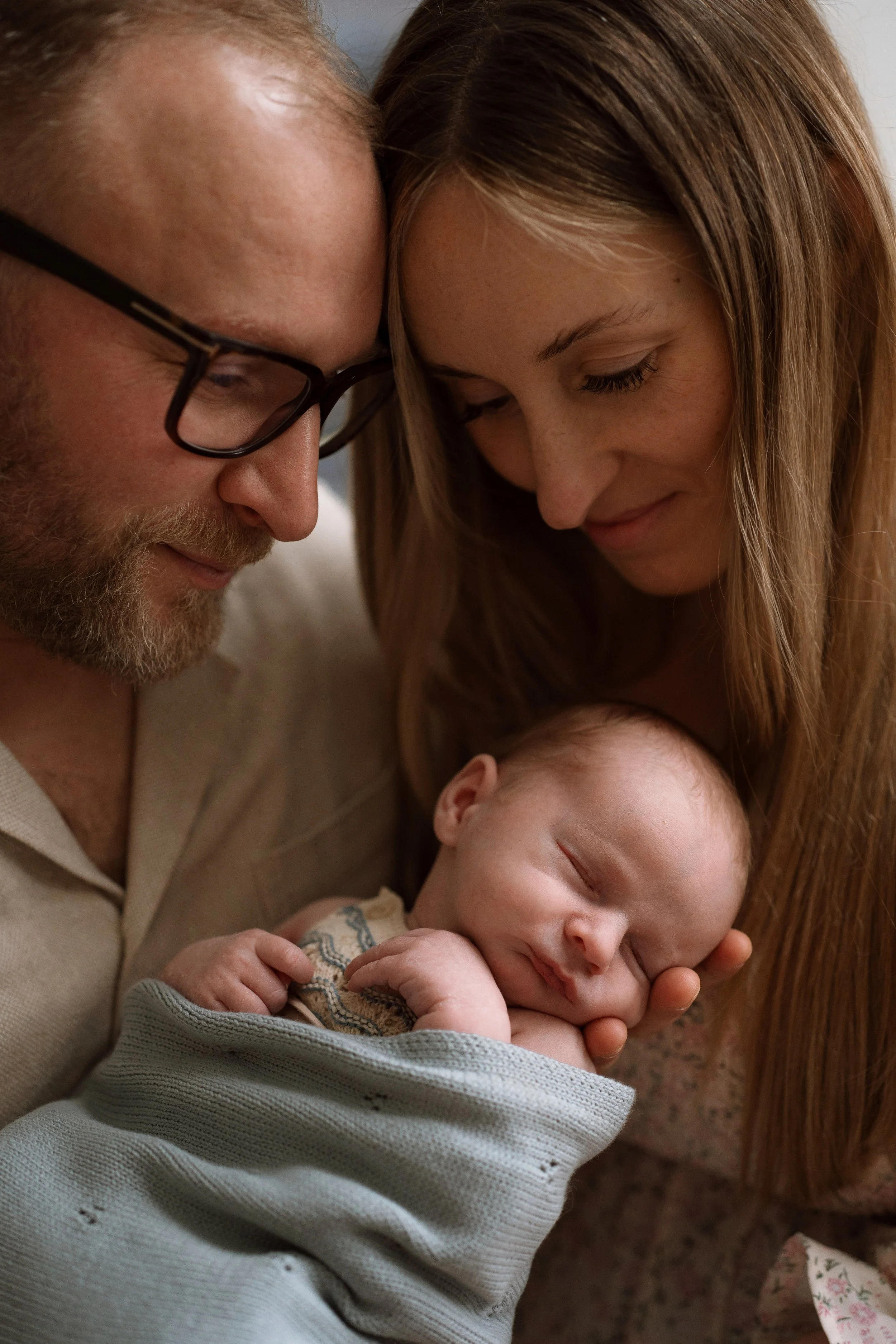 A family of three celebrating a new baby's arrival at home, with the father holding the sleeping infant and both parents smiling at the newborn.