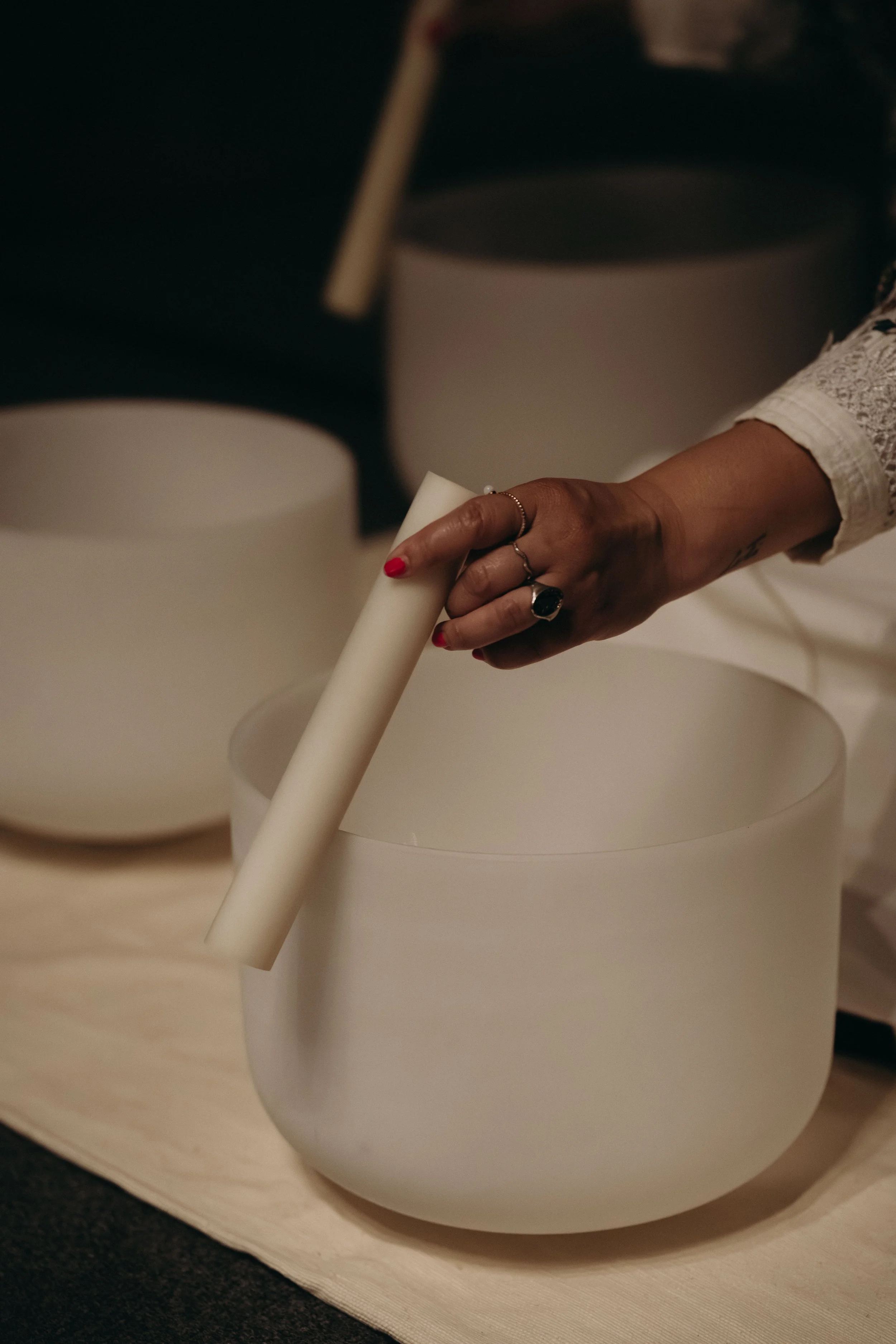 A person's hand with red nail polish and rings holding a white paper tube over a white vase or container, with another similar container or vase in the background, on a light-colored surface.
