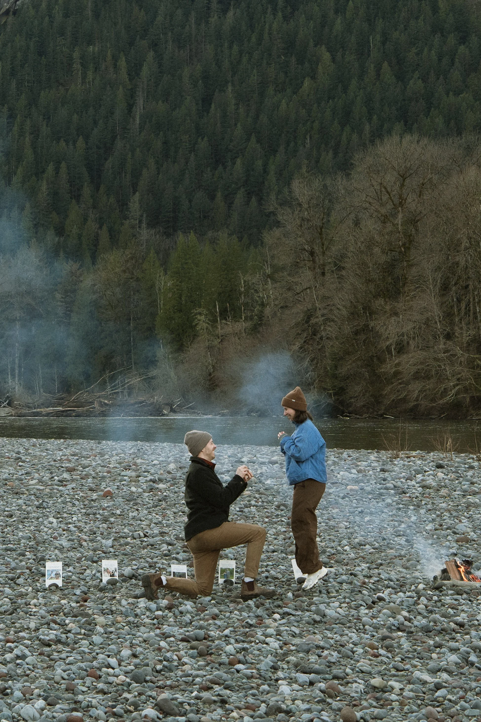Two people are on a rocky riverbank during sunset or sunrise. One is kneeling and proposing marriage to the other, who is standing with a smile. The man kneeling is holding a ring, and there are small signs or displays on the ground nearby. Smoke or 
