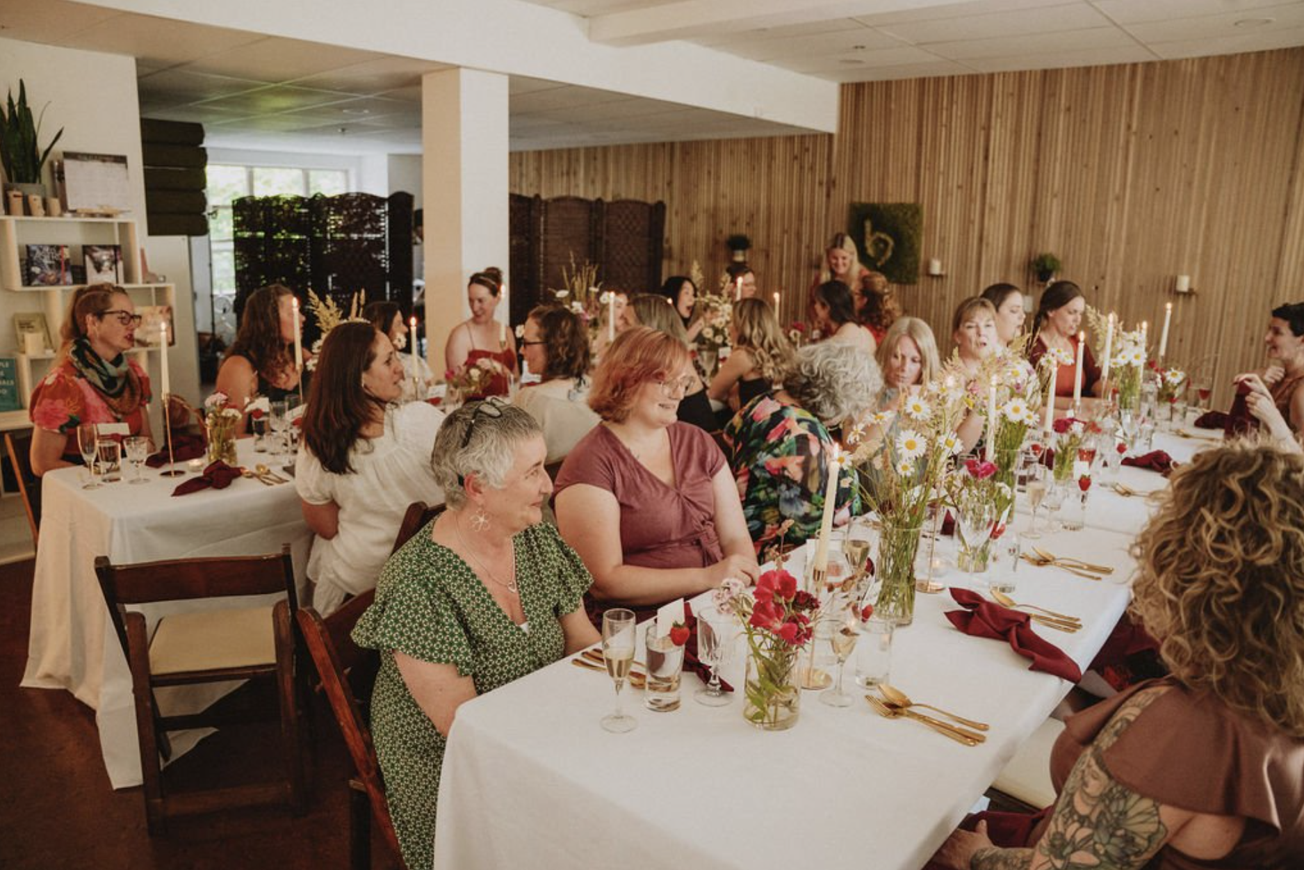 A group of women sitting at a long dining table decorated with flowers, candles, and elegant tableware, attending a social gathering in a warmly lit room with wooden walls.