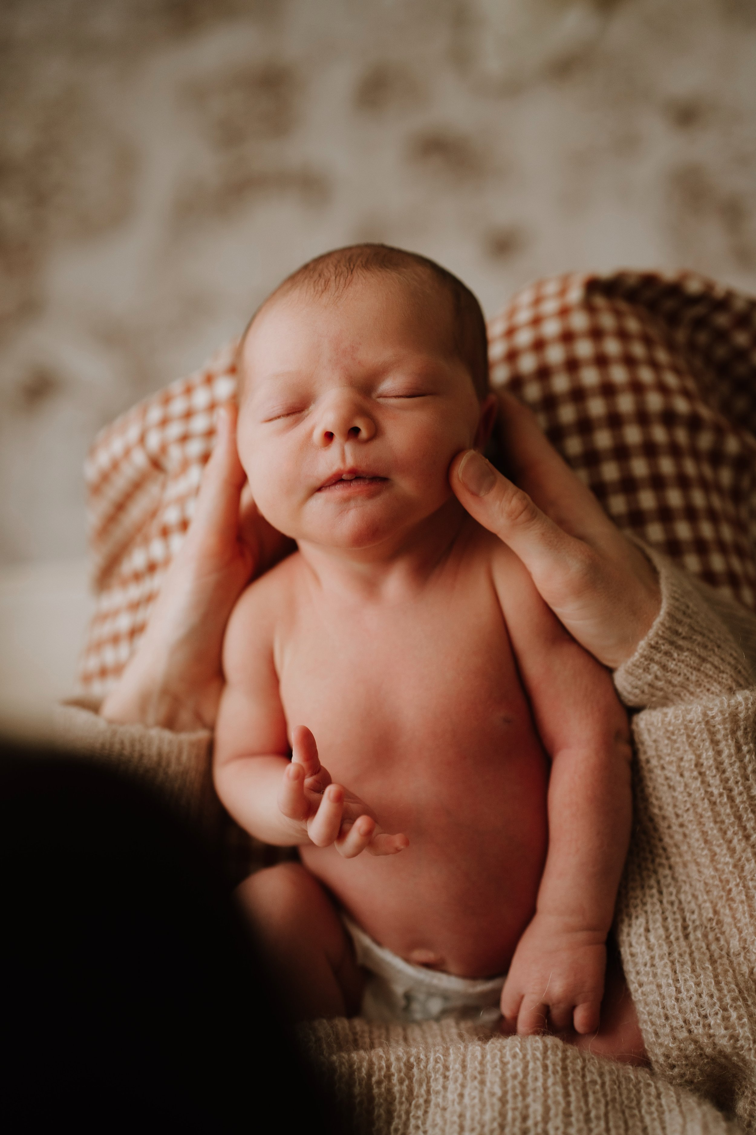 Close-up of a newborn baby with closed eyes, supported by hands, with a beige knitted blanket and a checkered cloth in the background.
