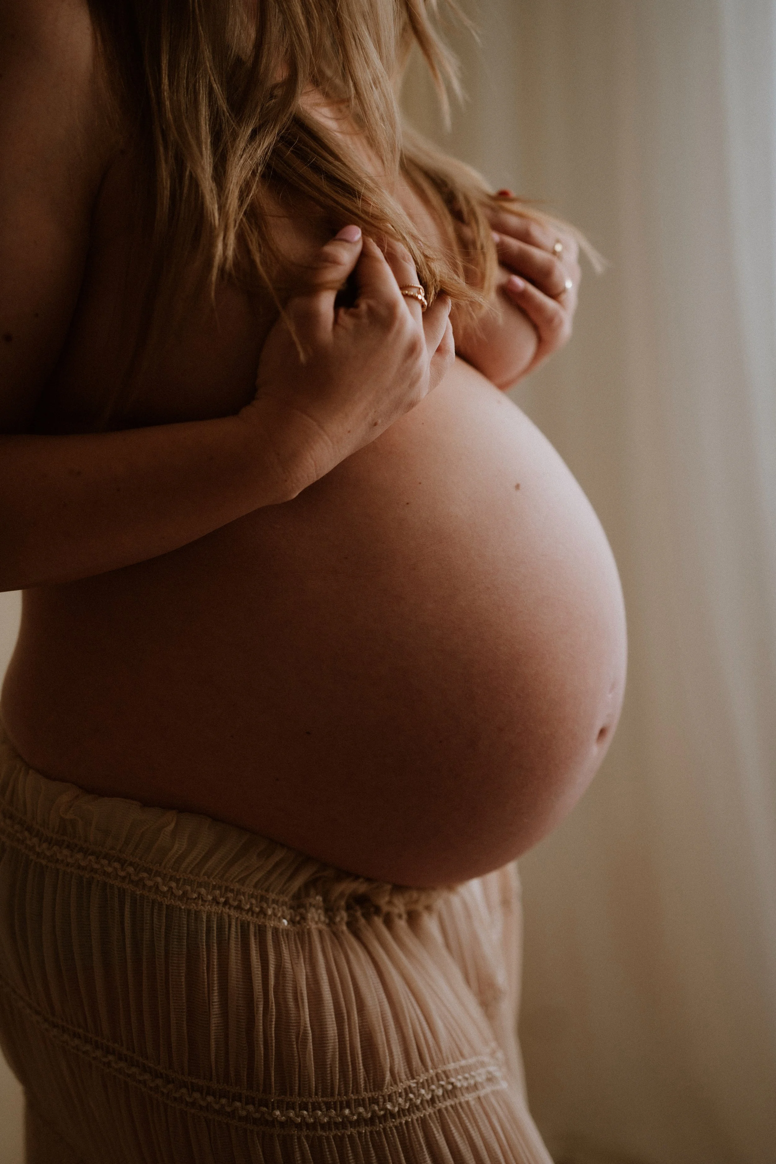 A close-up of a pregnant woman gently touching her chest with her hands, standing indoors with soft natural lighting, wearing beige pleated pants.