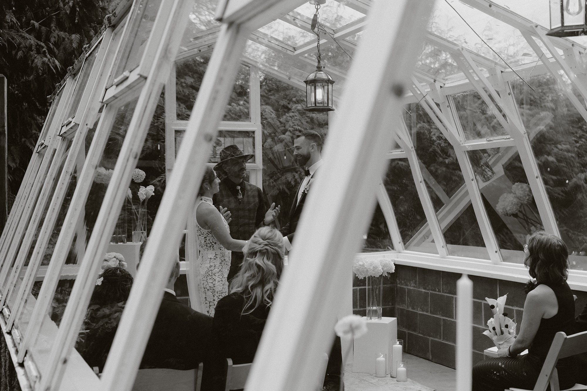 A black and white photo of a wedding ceremony inside a glass greenhouse, with a bride and groom holding hands and facing each other, surrounded by guests, with candles and floral decorations.