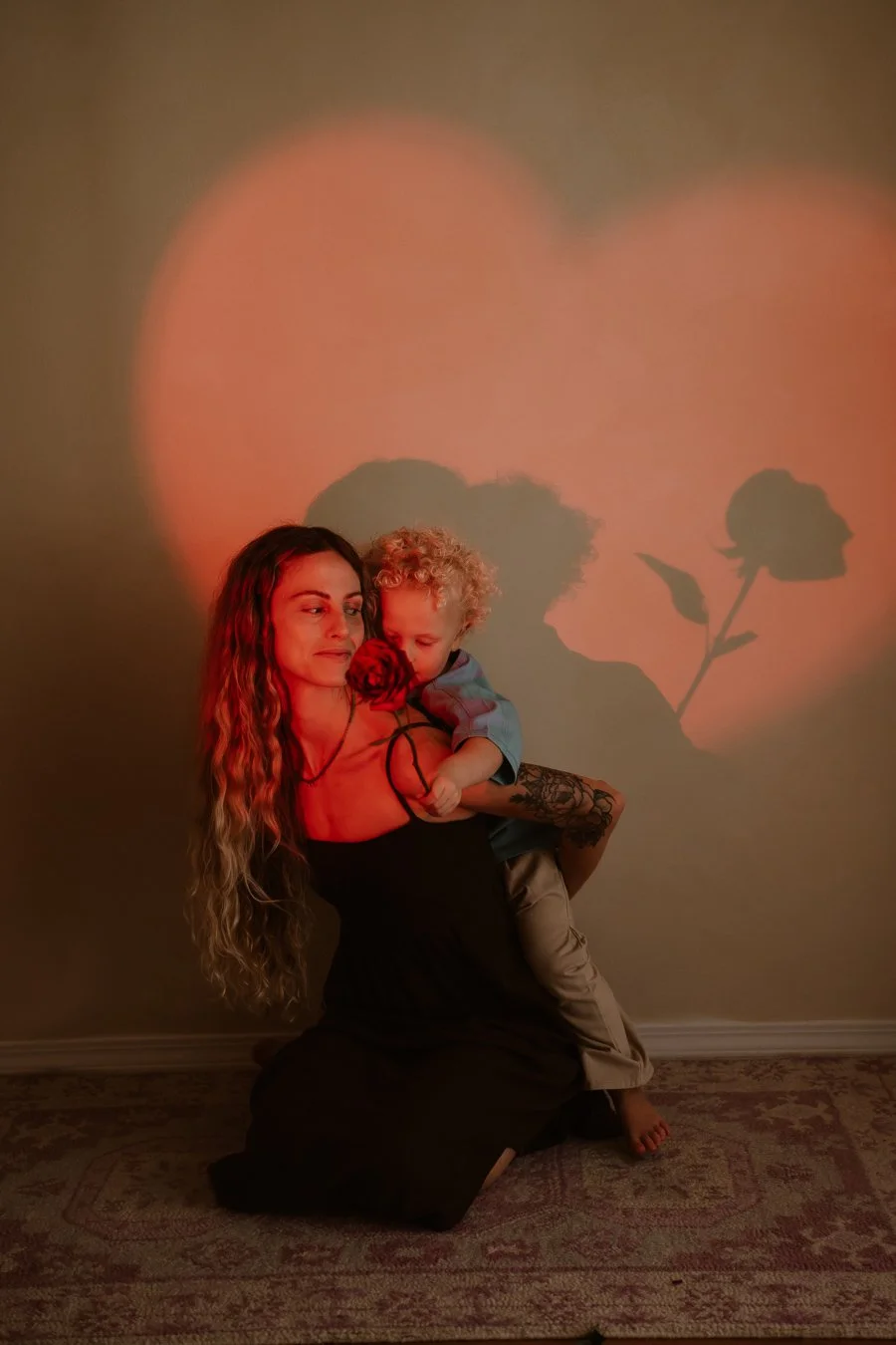 A woman with long curly hair and a tattoo on her arm is kneeling on a carpeted floor, holding a young boy with curly blonde hair. The boy is hugging a red rose and resting his head on her shoulder. A large red heart with a rose silhouette is projecte
