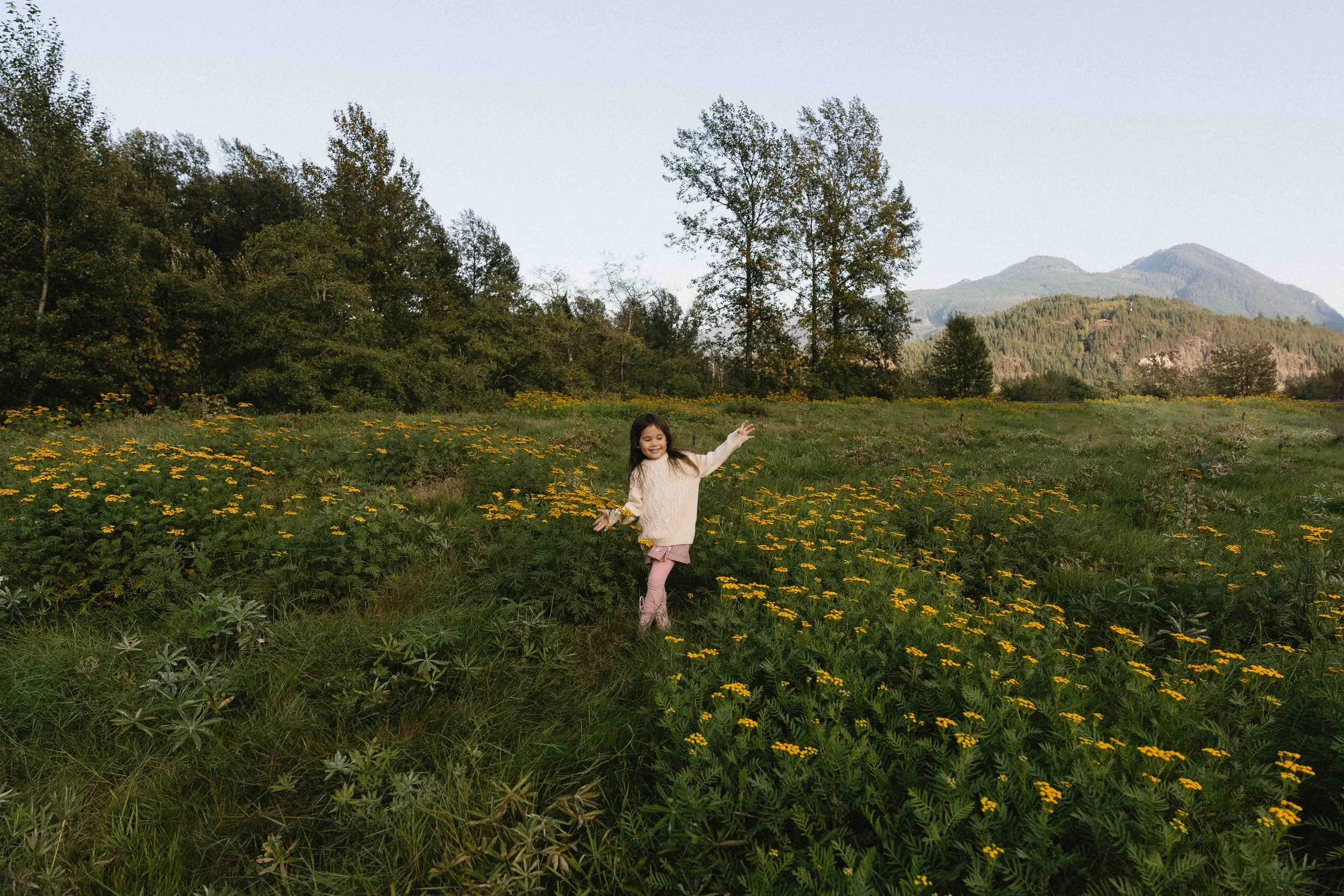 a young girl are in a field of white flowers, with trees and mountains in the background. The woman is kneeling and holding the girl’s hands while smiling at her.