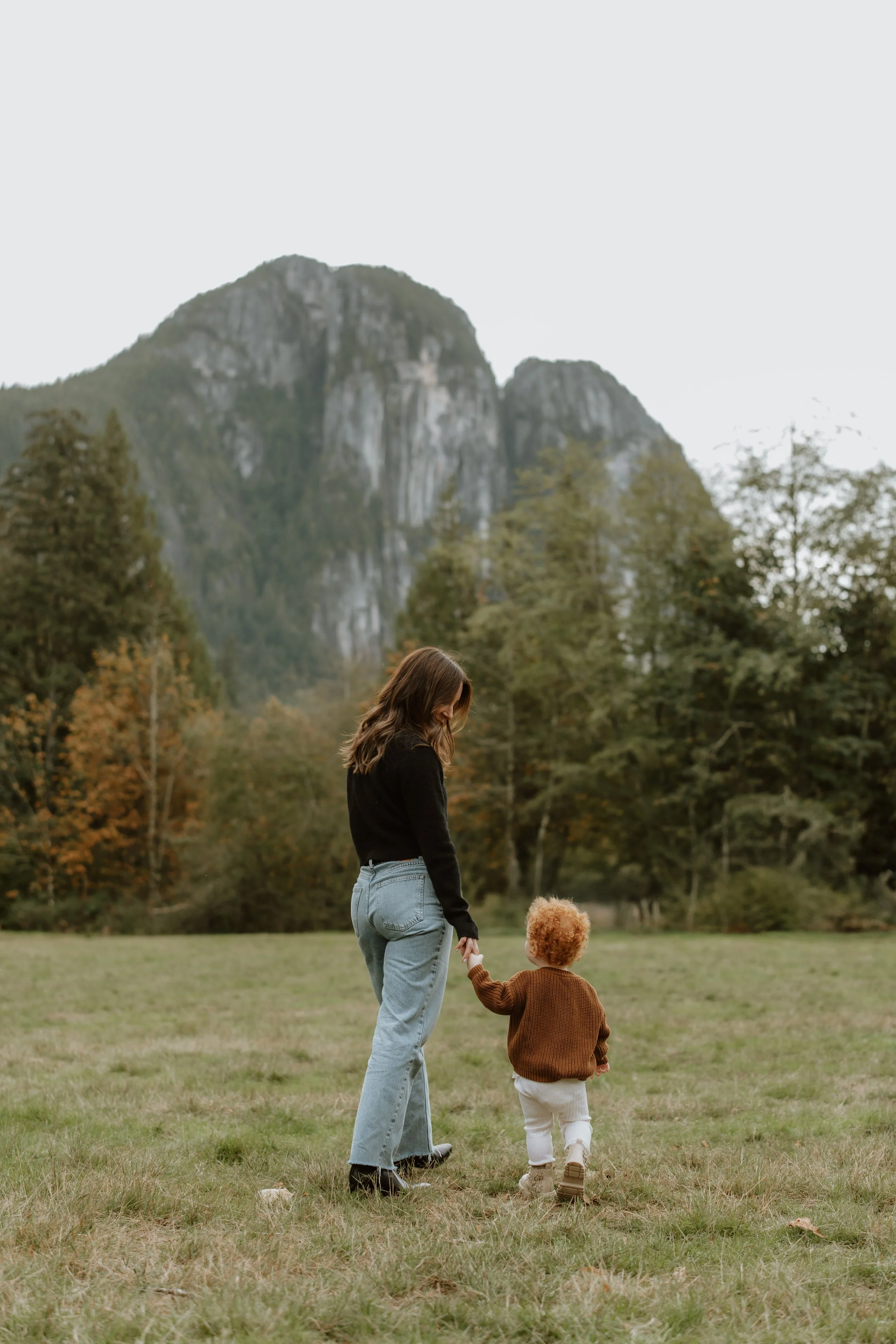A woman and a young child walking hand in hand across a grassy field with trees and mountains in the background.