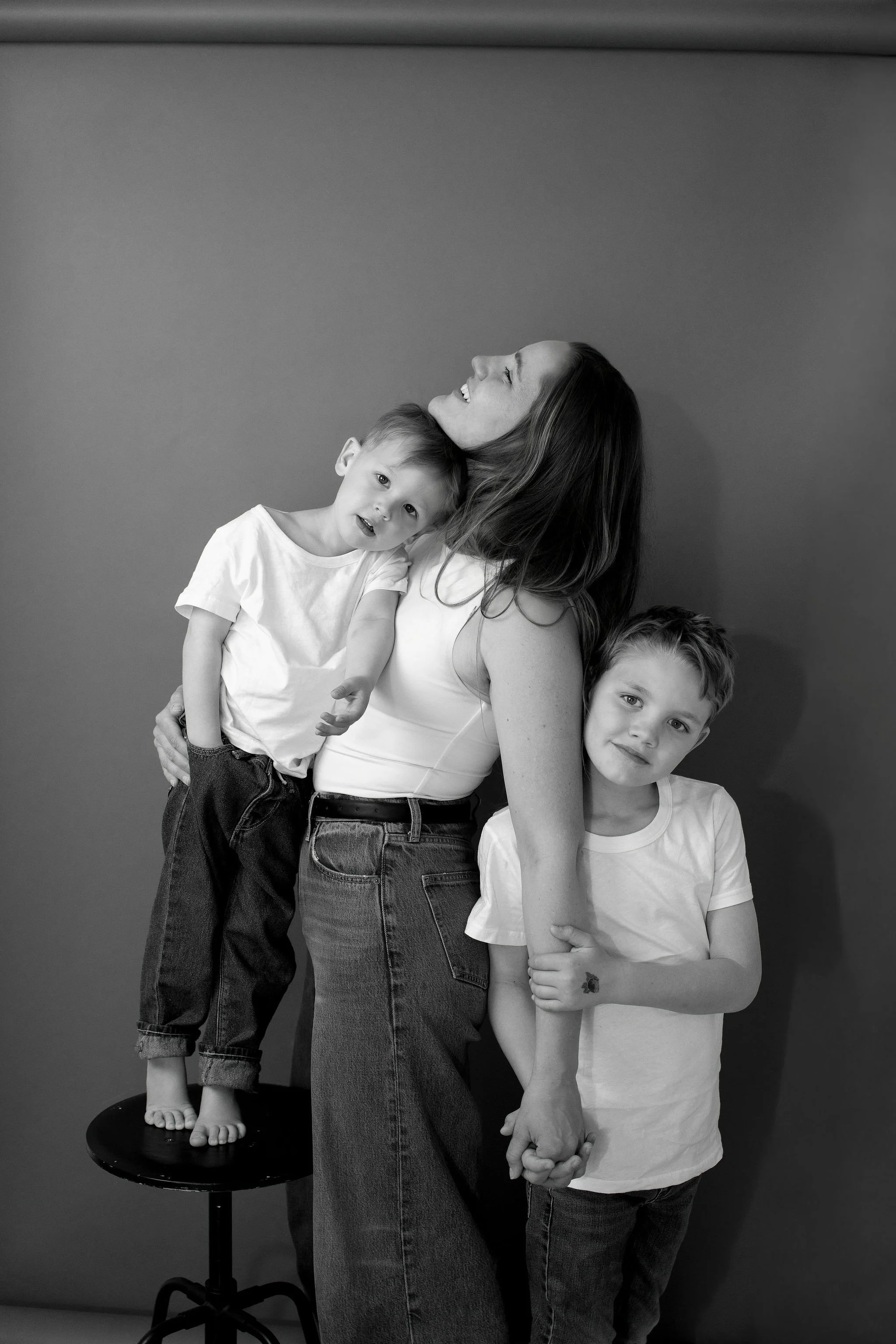 A woman with long hair smiling while holding a boy with short hair who is leaning on her shoulder. To her right, a girl with short hair is holding her arm and looking at the camera, and a boy with wavy hair is standing to the side, also looking at th