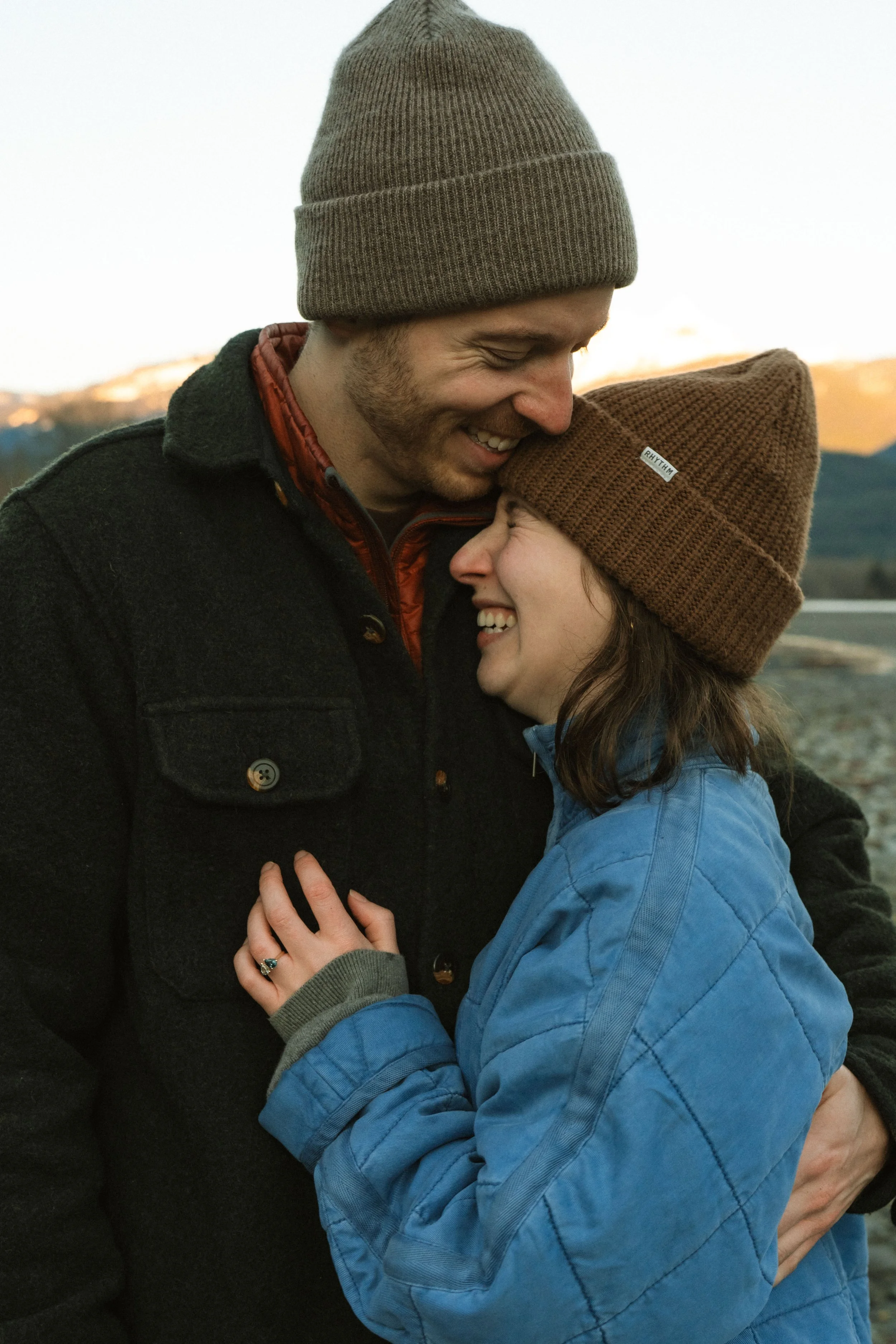 A man and woman sharing a joyful moment outdoors, wearing warm clothing including beanies, with their foreheads touching and smiling.