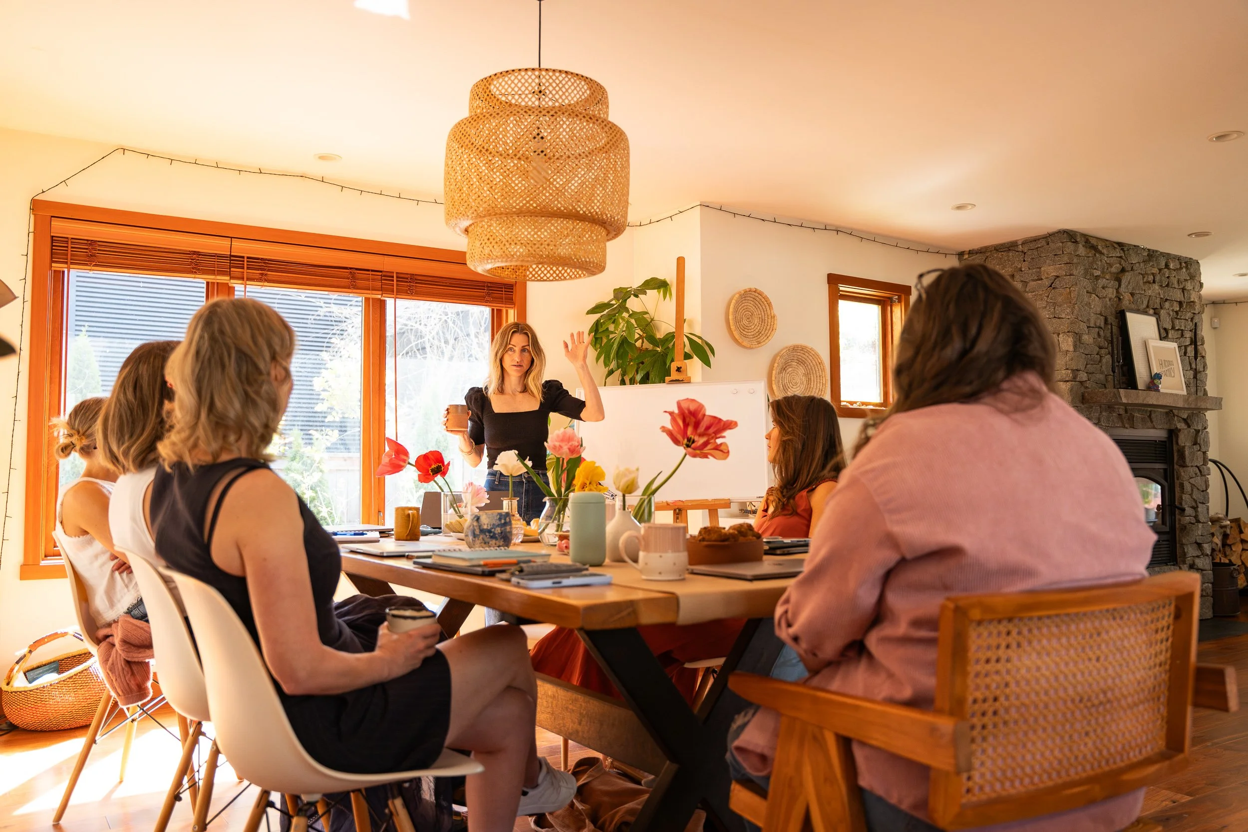 A woman standing at the front of a room giving a presentation to a group of women seated at a dining table with flowers, notebooks, and coffee cups. The room has large windows, wooden blinds, and a stone fireplace.