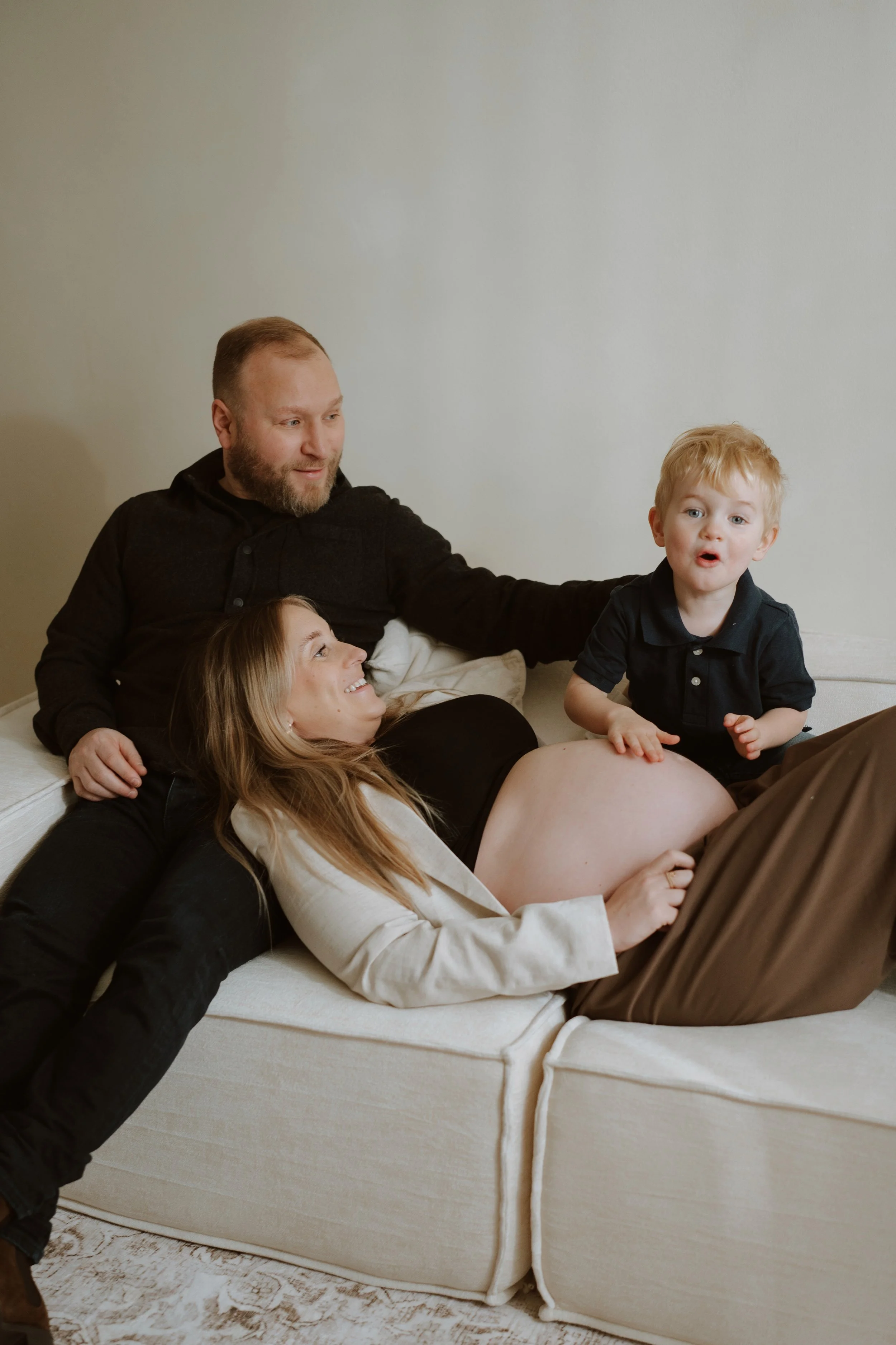 A group of four people, including two children, are sitting on a beige sofa in a room with a plain light-colored wall. They appear to be engaged in a playful and relaxed moment.