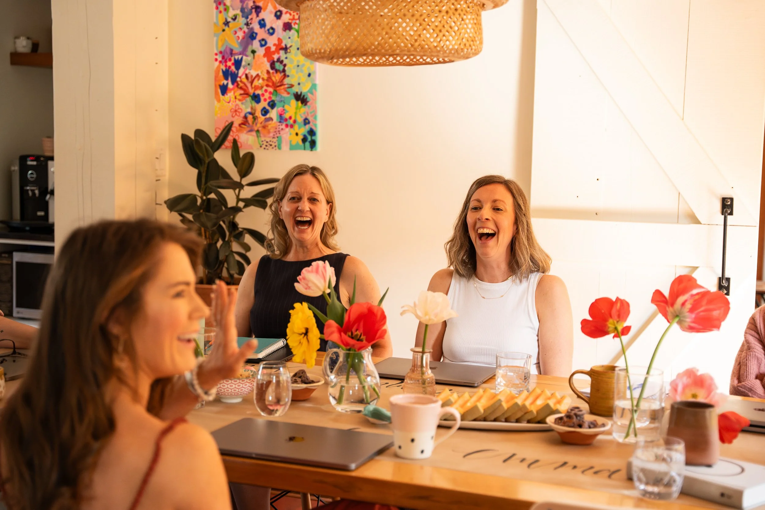 Four women sitting at a wooden table, laughing and enjoying each other's company during a gathering. The table is decorated with colorful flowers, and there are drinks, snacks, and laptops on the table. The room has a cozy and cheerful atmosphere wit