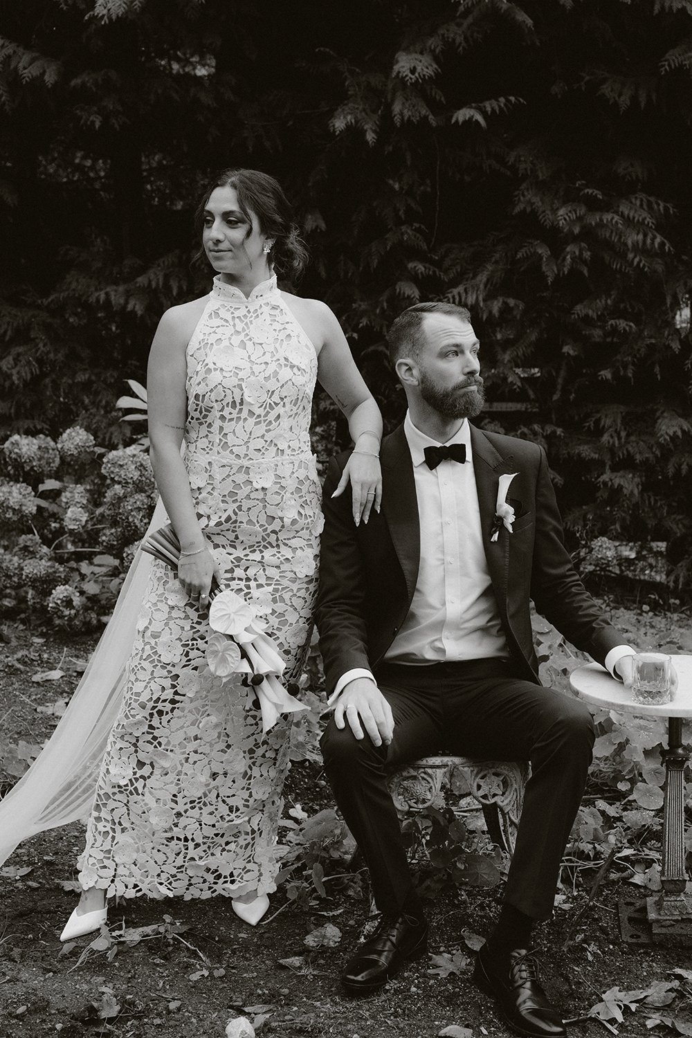 A black and white photograph of a woman in a lace dress and a man in a tuxedo, seated outdoors with trees and flowers in the background. The woman is standing with one hand on the man's shoulder, holding a bouquet, while the man is sitting on a chair