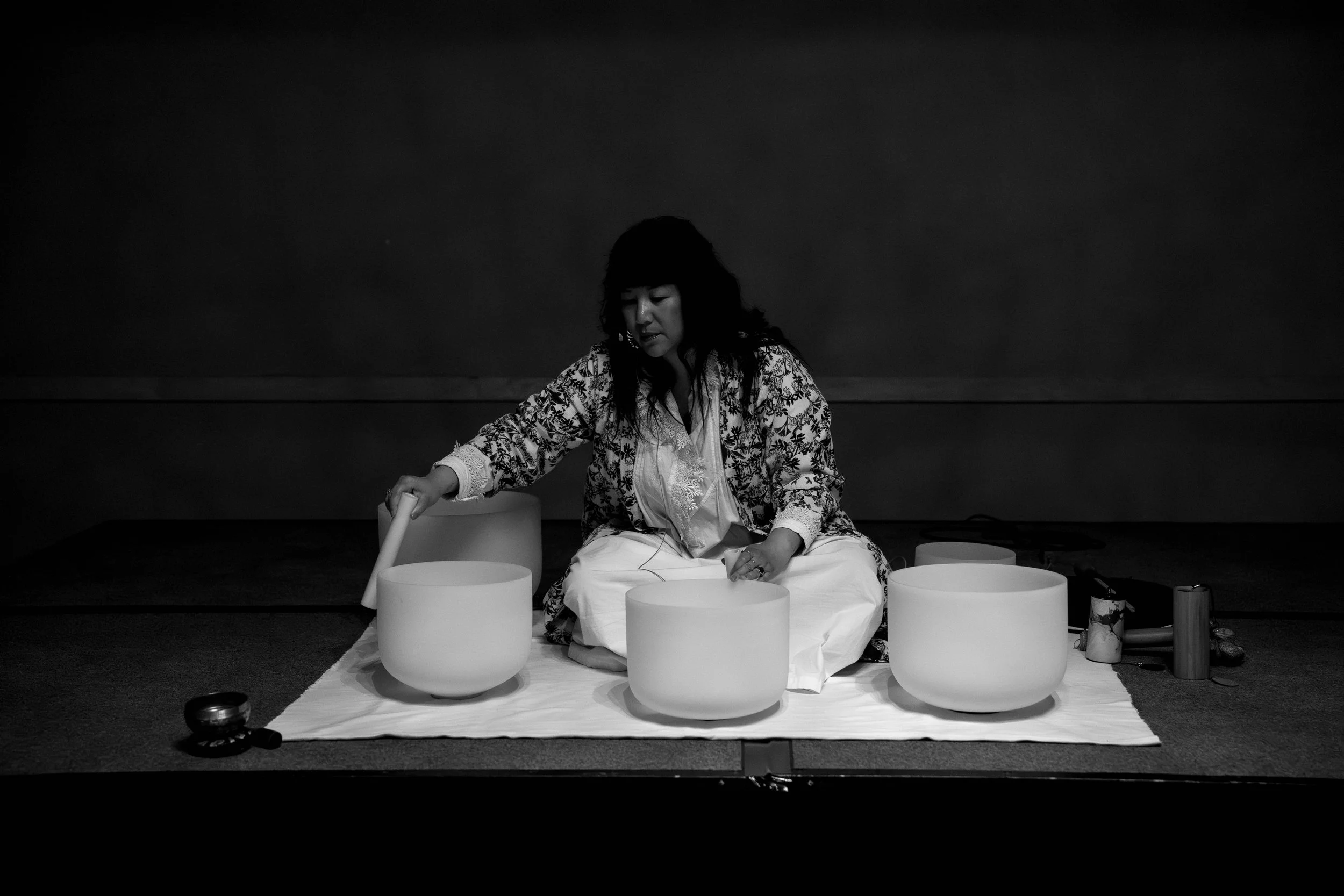 A woman wearing a patterned kimono sitting on a blanket with four large singing bowls in front of her, practicing sound therapy in a dark room.