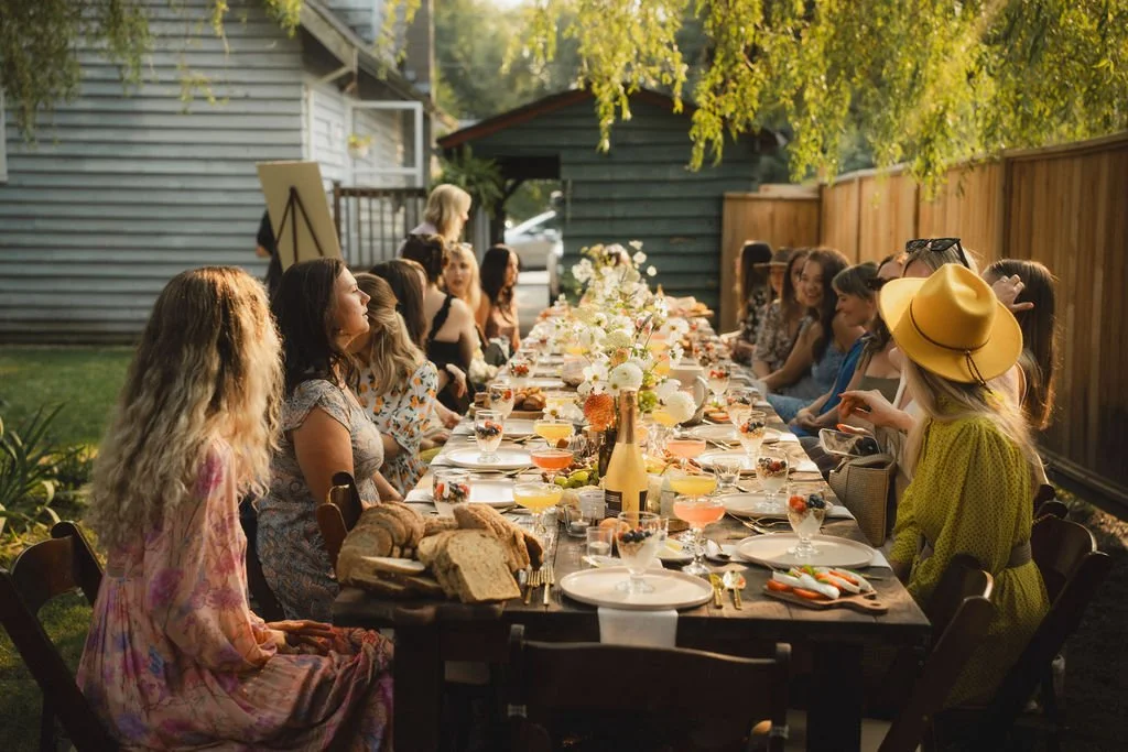 Outdoor dinner party with women sitting at a long table adorned with flowers, bread, and drinks, under a tree in someone's backyard during daytime.