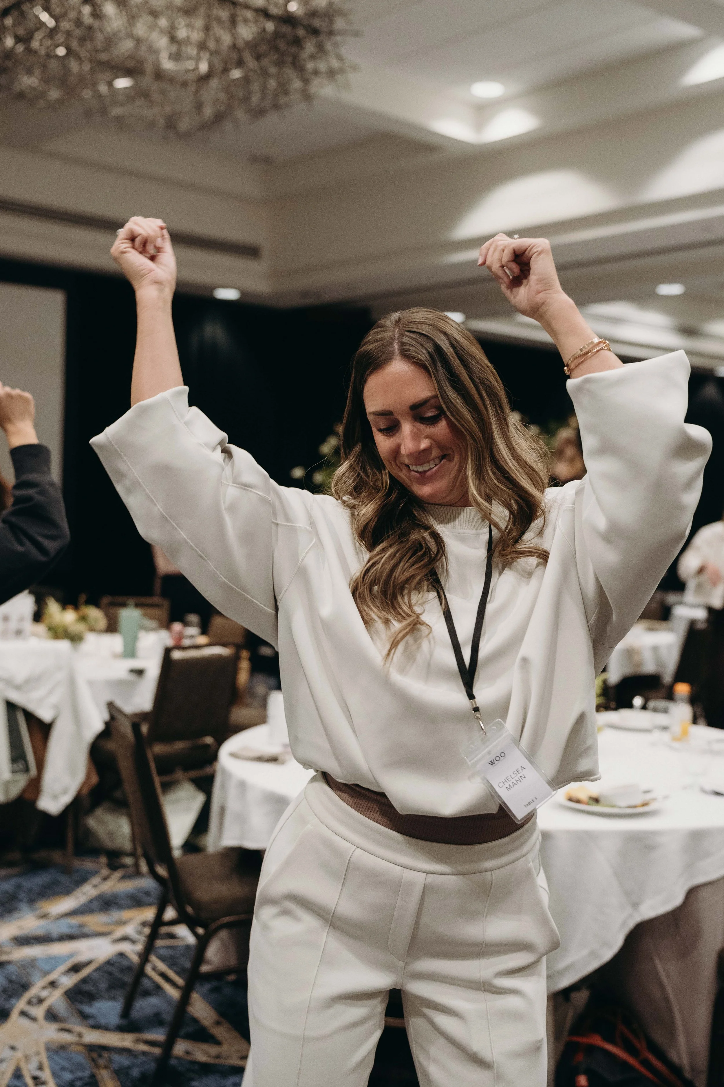 A woman with long, wavy hair dancing with her arms raised in a conference or event room with banquet tables in the background.