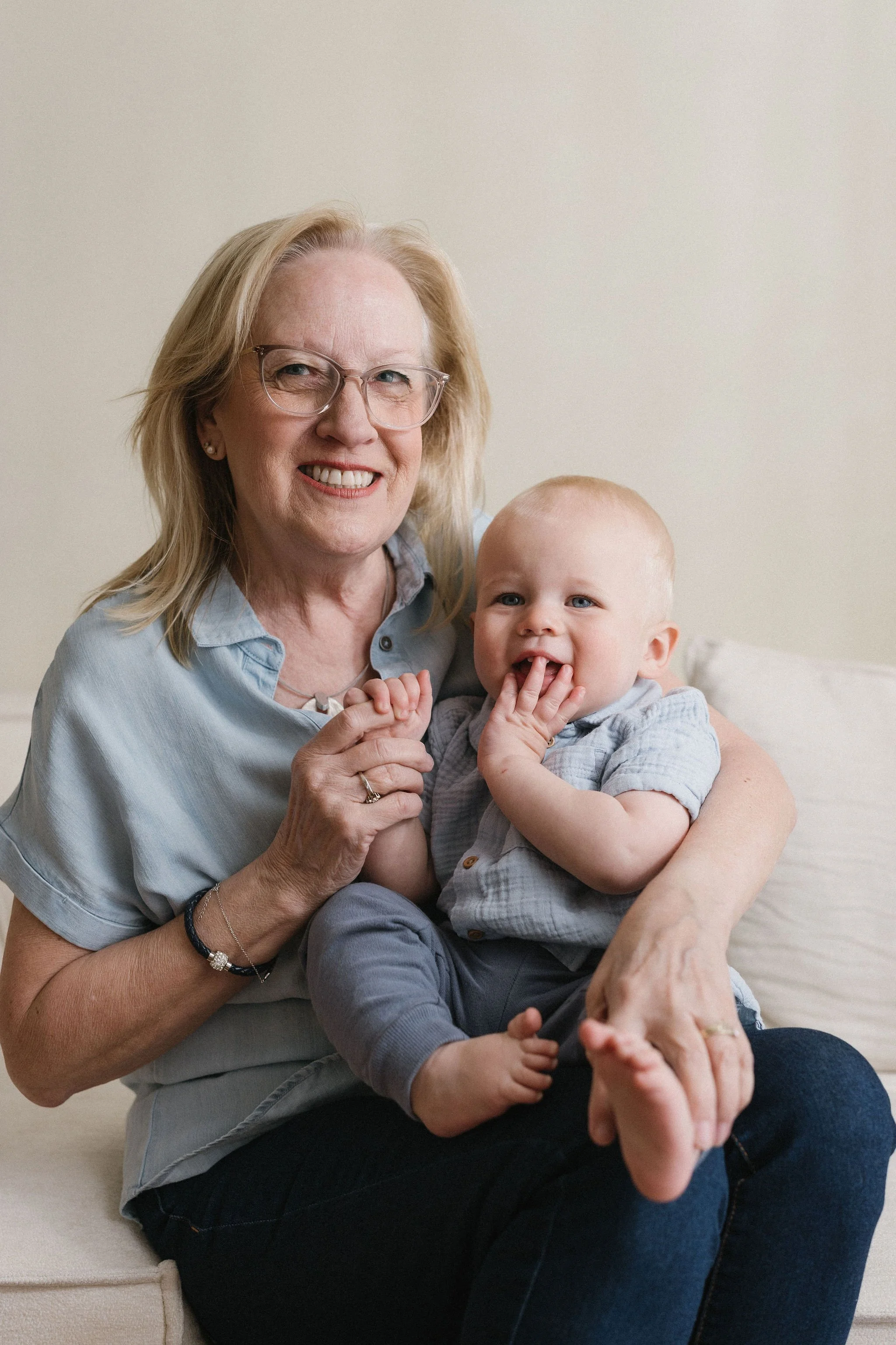 An elderly woman with glasses and a young child smiling and sitting on her lap, both holding hands and looking at the camera against a plain background.