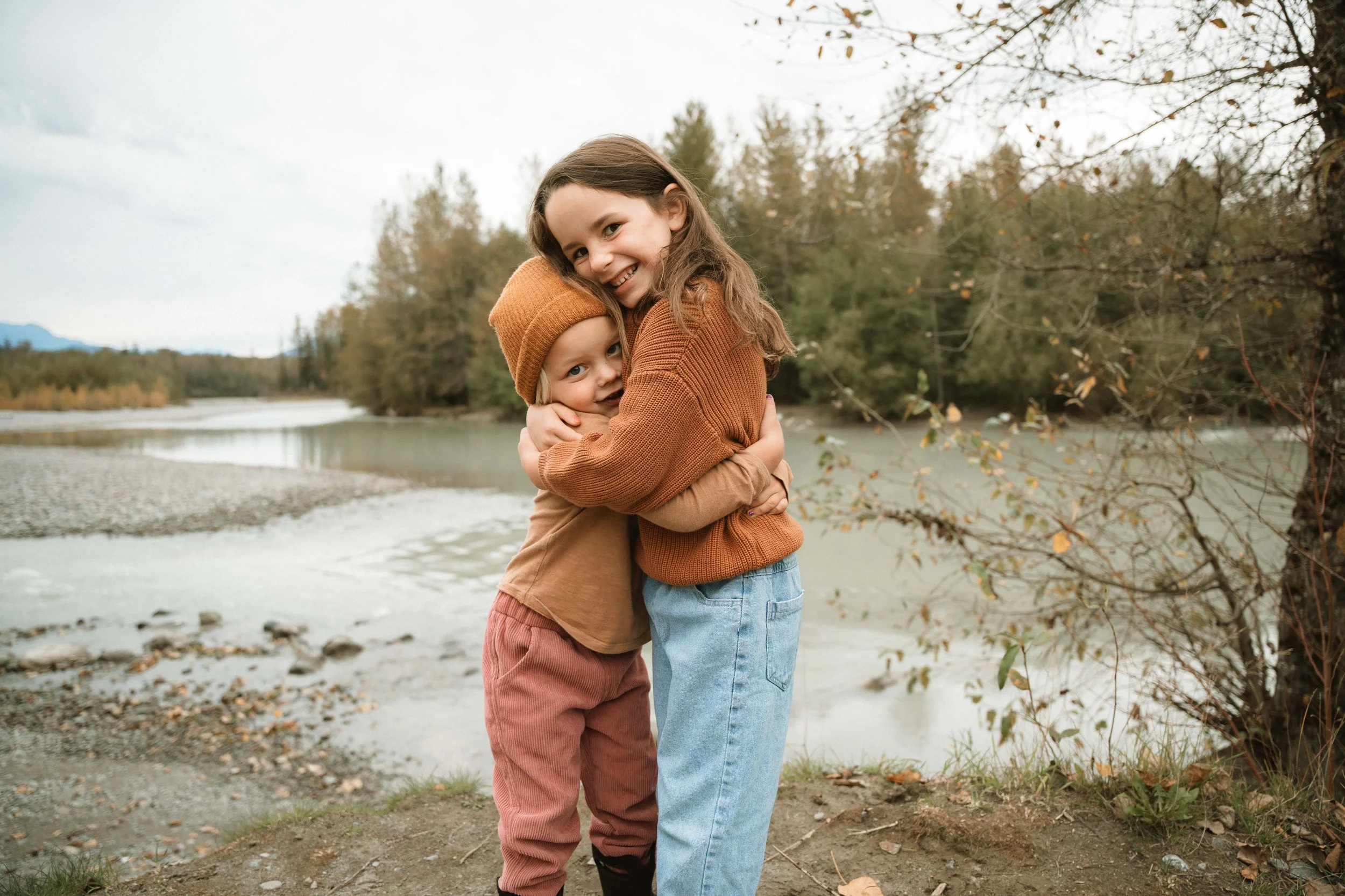 Two children, a girl and a boy, hugging outdoors near a river with trees in the background during fall.