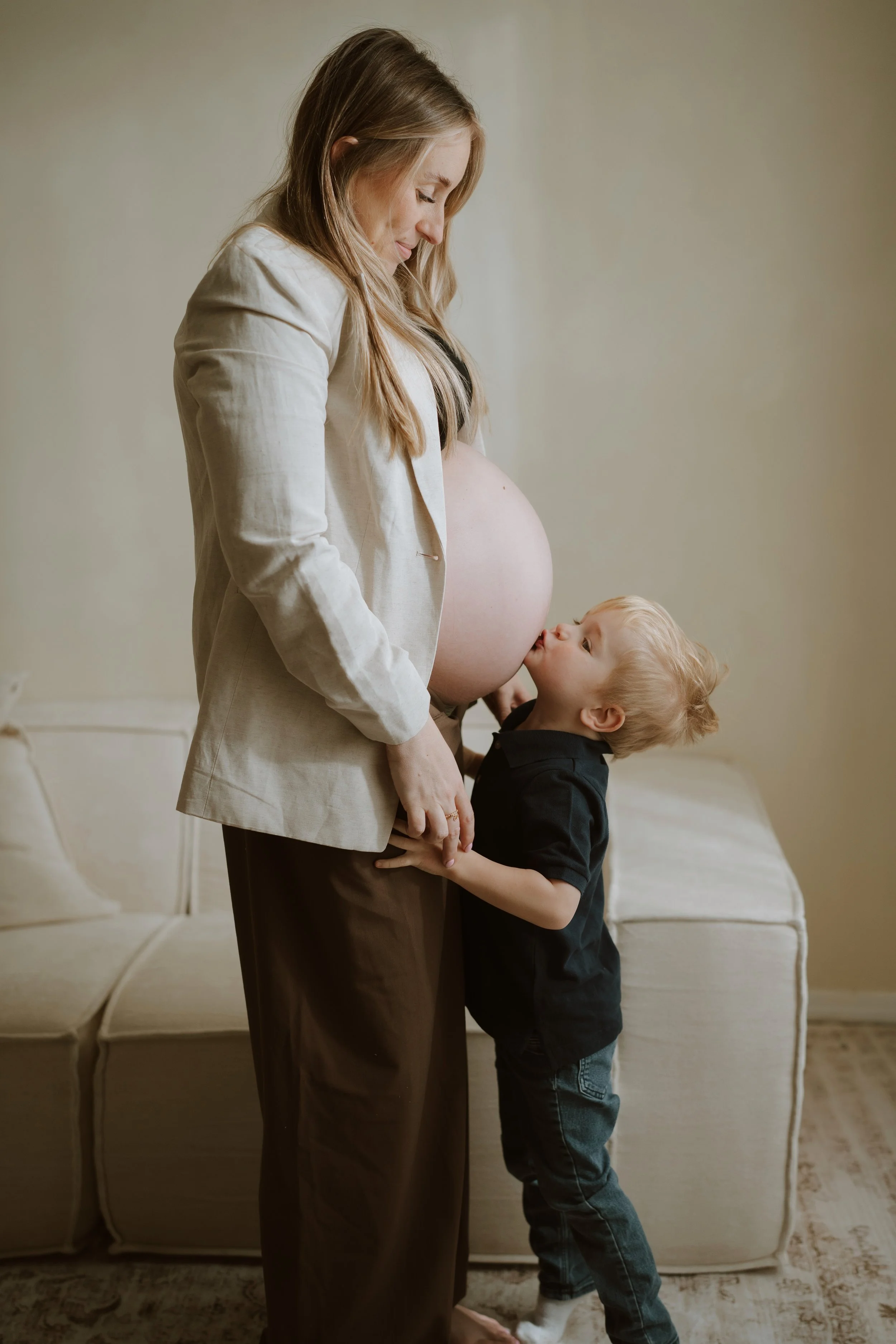 A pregnant woman with long blonde hair standing in a living room, looking down and smiling at a young boy who is kissing her pregnant belly. The woman is wearing a white blazer and dark pants, and the boy has curly blonde hair and is dressed in a bla