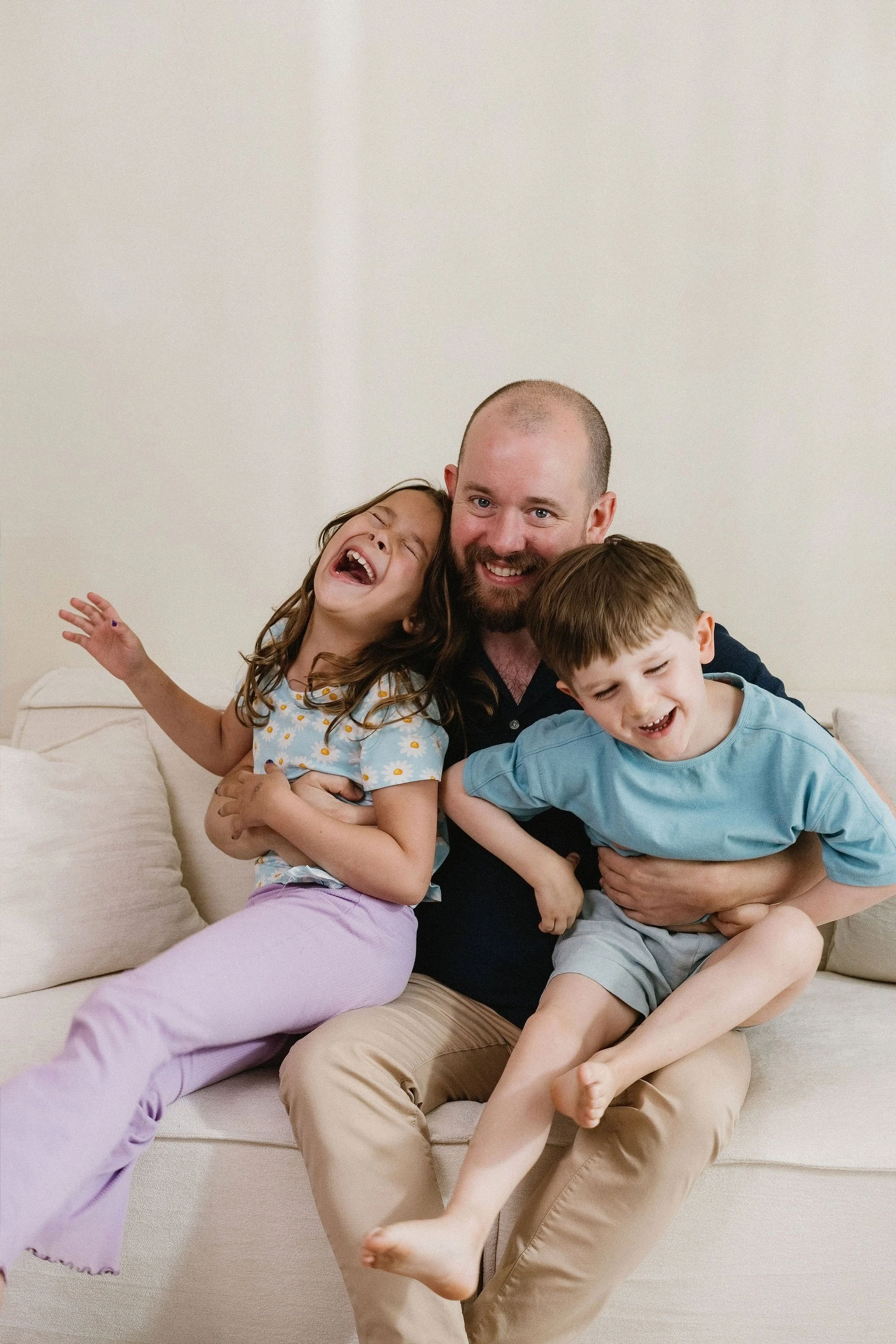 A joyful man with a beard and two children, a girl and a boy, sitting on a beige sofa against a plain white wall, laughing and embracing each other, in a cozy home setting.