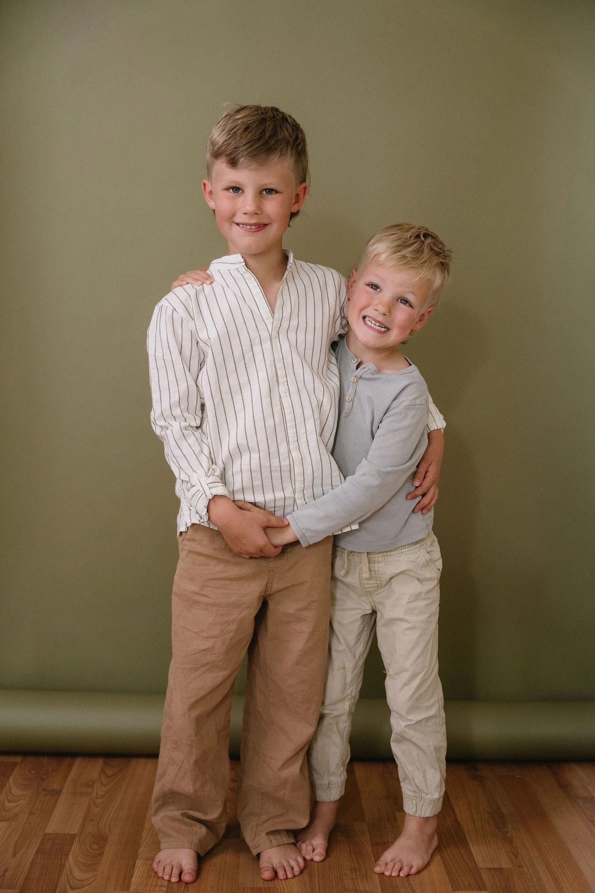 Two young boys standing close together indoors in front of a green wall, with their arms around each other, smiling and looking at the camera. The older boy is wearing a cream and brown striped shirt, beige pants, while the younger boy is wearing a l