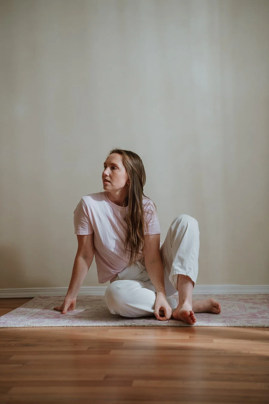 Woman sitting on a rug on hardwood floor, wearing a light pink t-shirt and white pants, looking to her left.