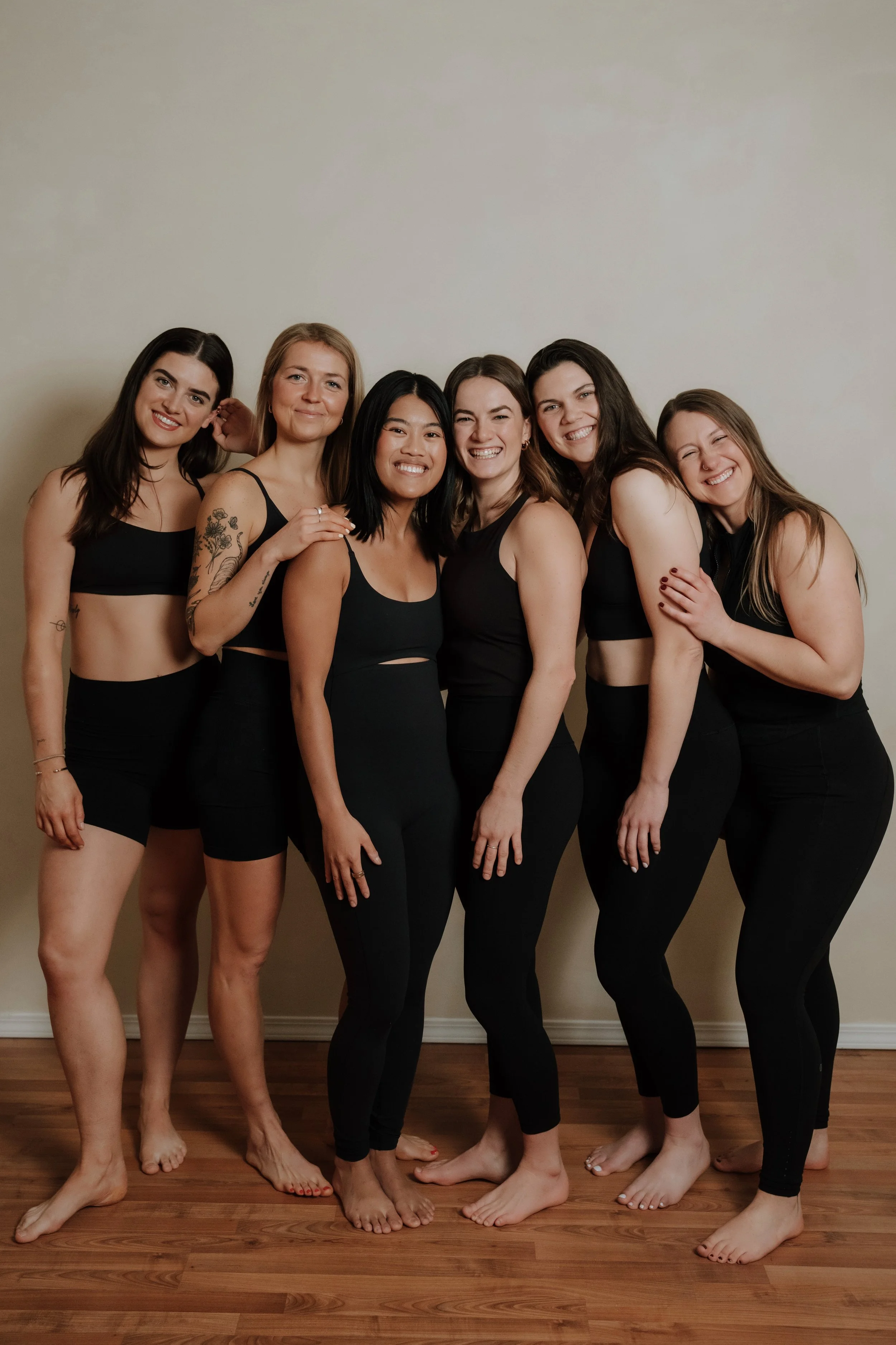 Six women standing close together indoors, smiling, dressed in black athletic wear, with some barefoot on a wooden floor against a plain wall.