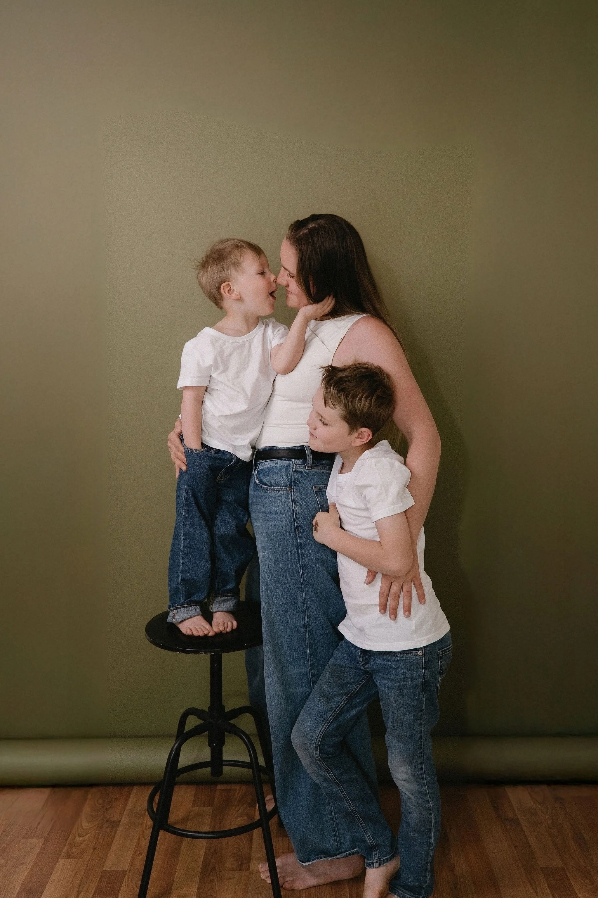 A woman with two young boys, one standing on a stool and the other standing on the floor, all wearing white shirts and blue jeans. The boy on the stool is touching the woman's face and leaning in for a kiss, while the woman and the other boy are smil