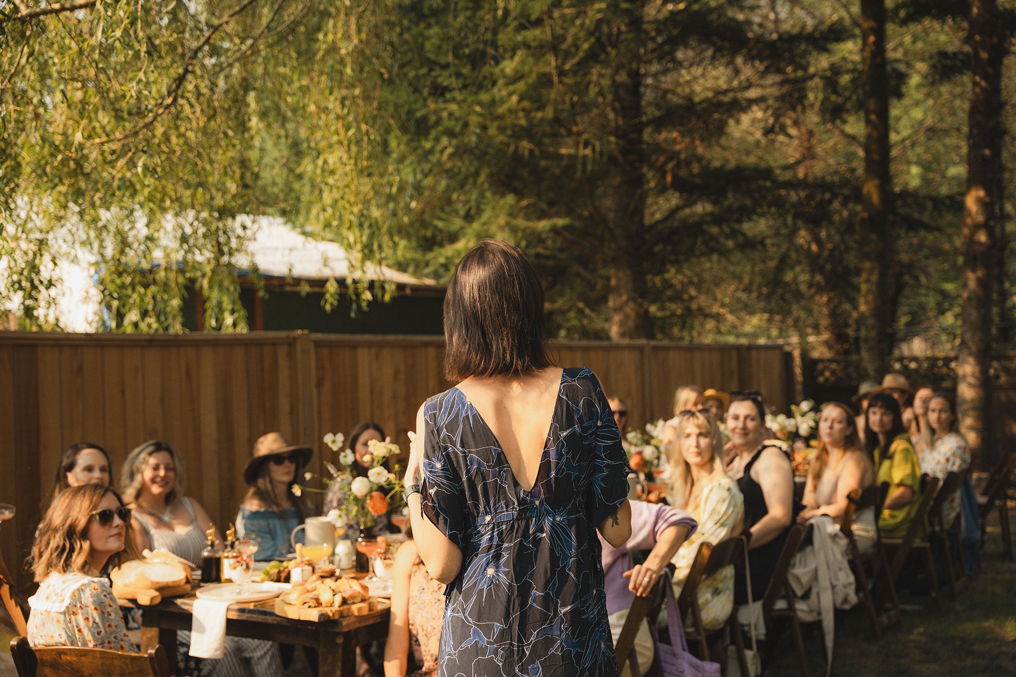Woman speaking to a group of people seated at an outdoor table decorated with flowers and food during a sunny day in a garden.