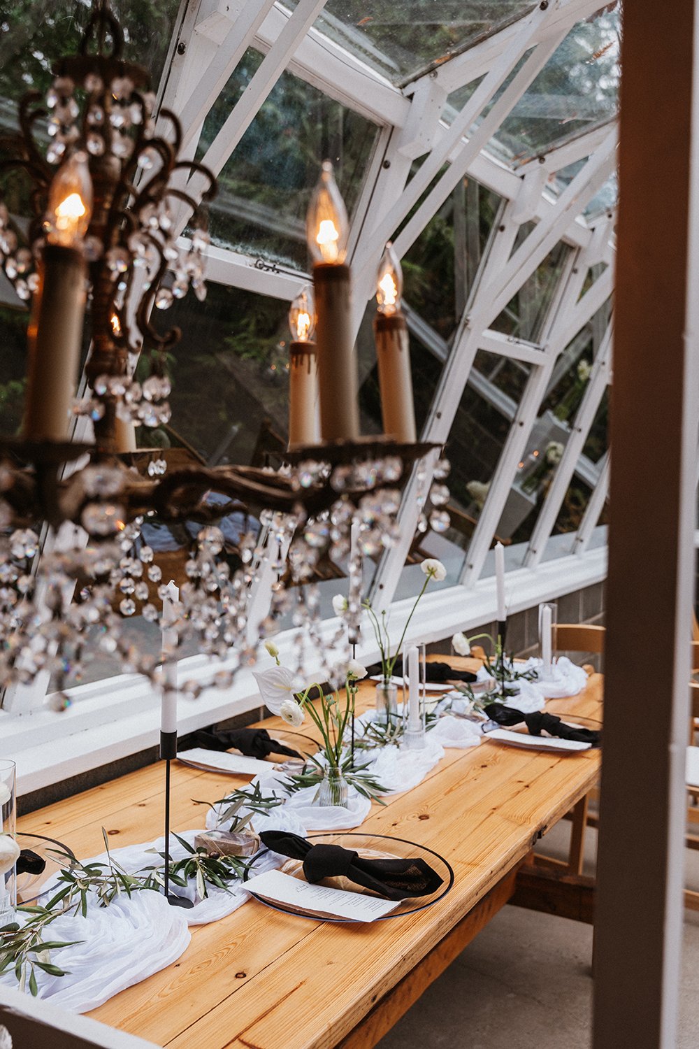 A decorated dining table with white tablecloth, black napkins, and floral centerpieces, inside a glass conservatory with a chandelier and large windows.
