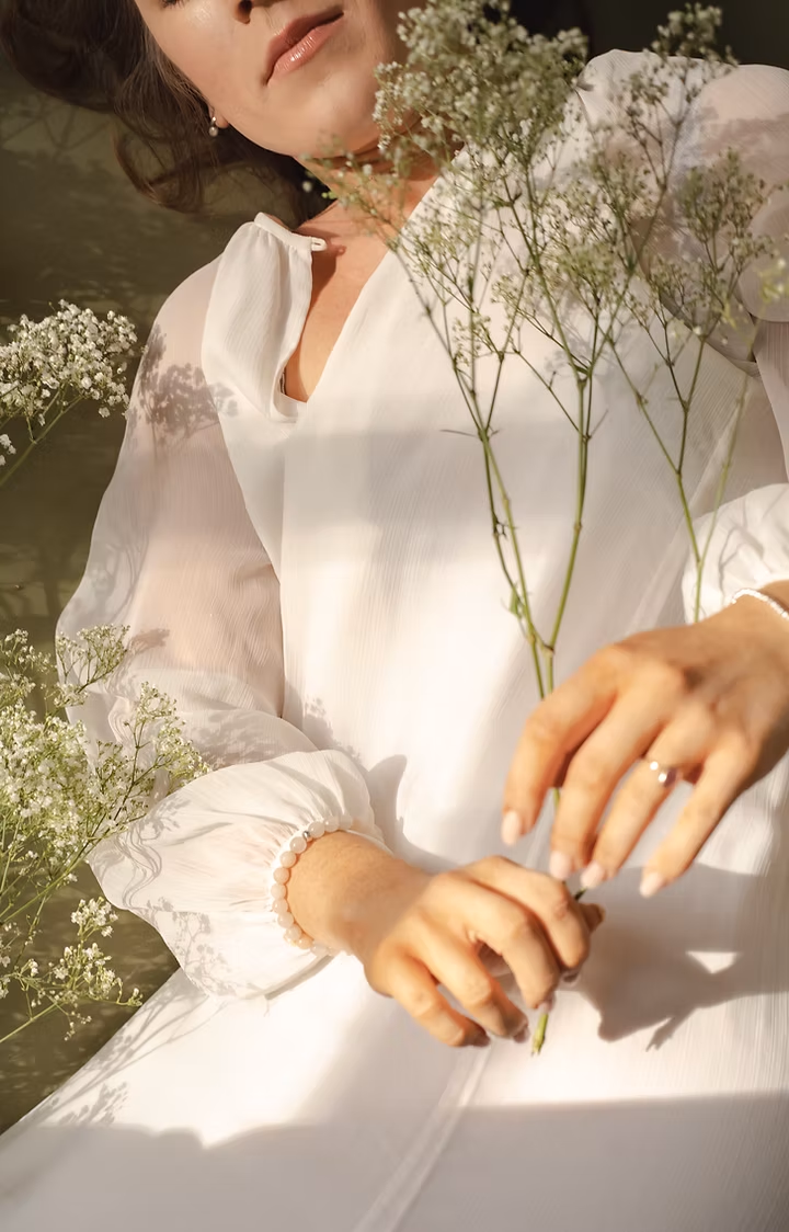 A woman in a white dress holding delicate white flowers, with a pearl bracelet on her wrist and ring on her finger, in soft natural lighting.