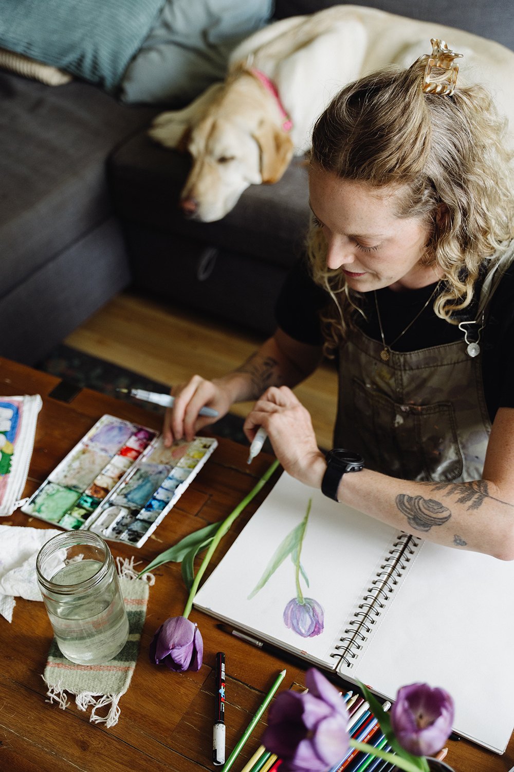 A woman with curly hair and tattoos on her arm is painting a purple tulip in a sketchbook, while a dog with a pink collar is sleeping on a couch in the background.
