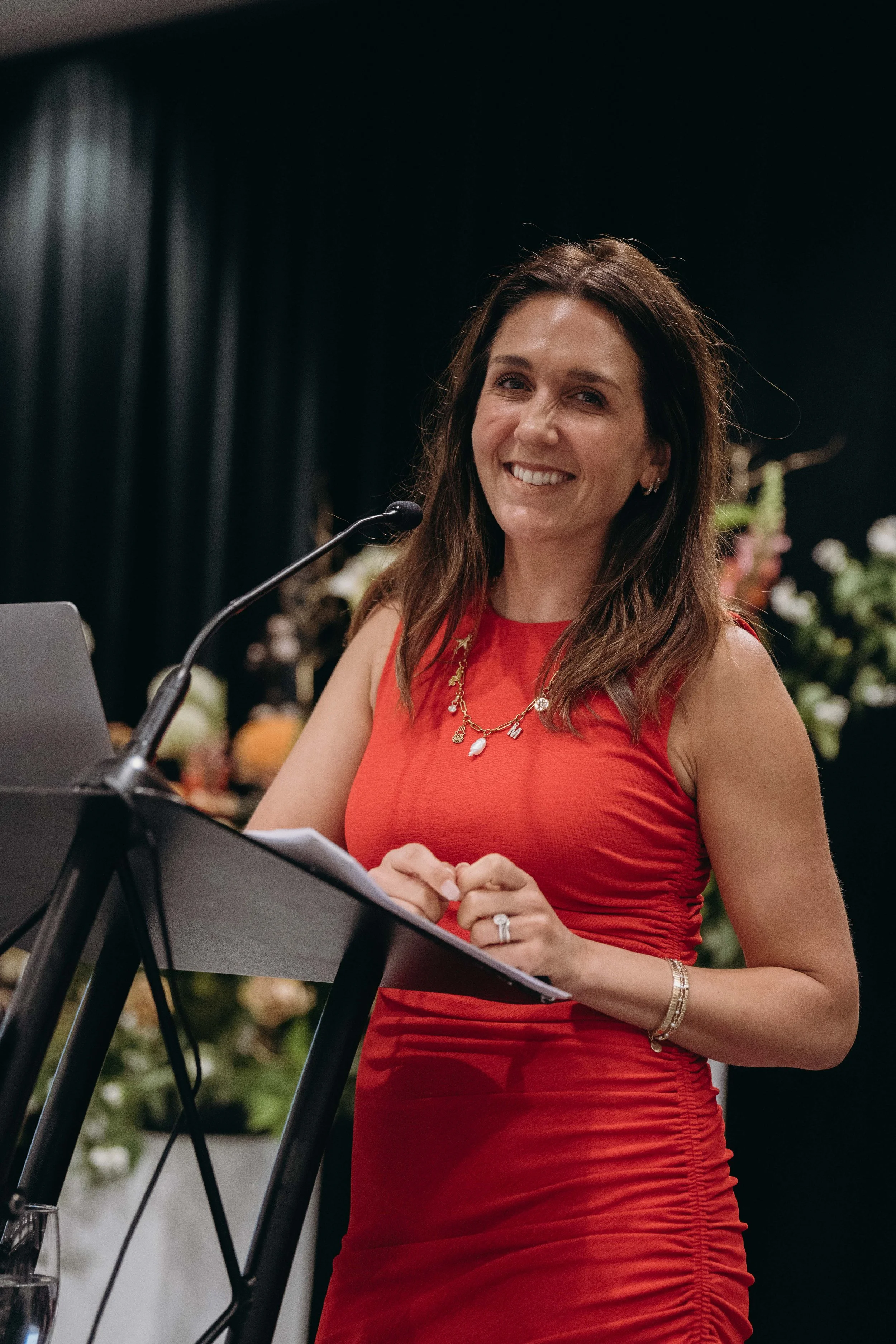 A woman with dark brown hair in a loose style, smiling, wearing a red sleeveless dress, a gold necklace, and bracelets, standing at a lectern with a microphone, indoors with floral arrangements in the background.