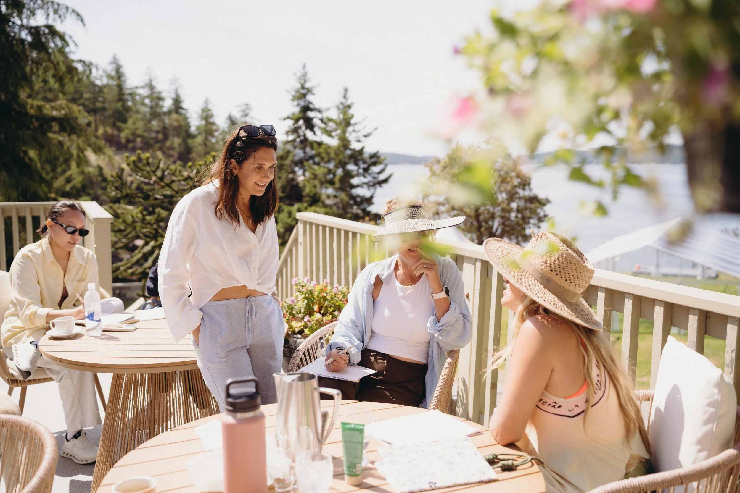 A group of women are gathered on a patio near a body of water, engaging in conversation on a sunny day. Some women are wearing hats and sunglasses, with a table set with drinks and snacks in front of them.