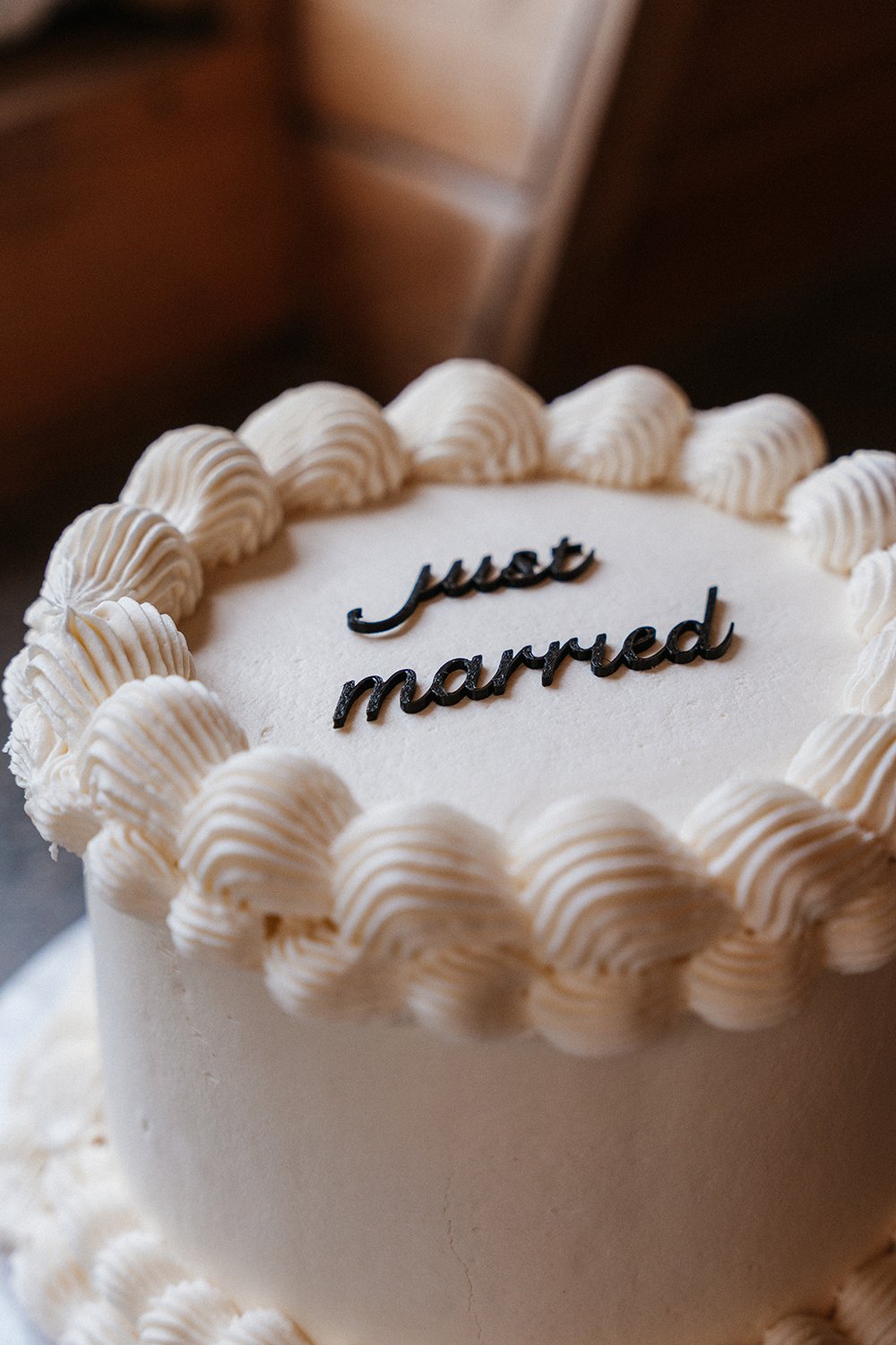 A wedding cake with white frosting, decorated with piped cream swirls around the top edge. The cake has a message that reads 'just married' in black cursive icing.