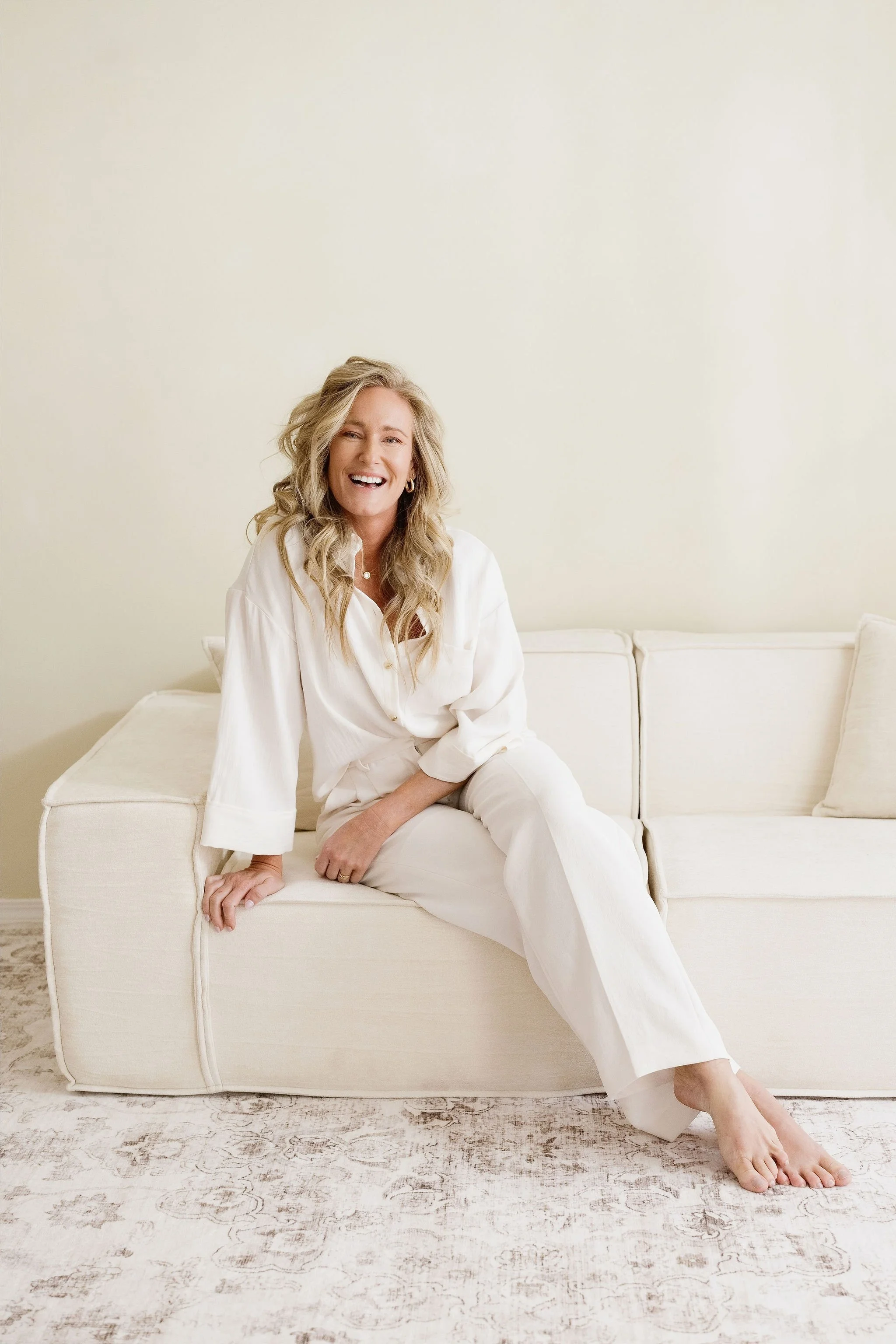 A woman with blonde wavy hair, wearing a white blouse and white pants, sitting on a cream-colored couch. She is smiling and laughing, with one leg crossed over the other, barefoot, in a neutral room with a light-colored wall and patterned rug.