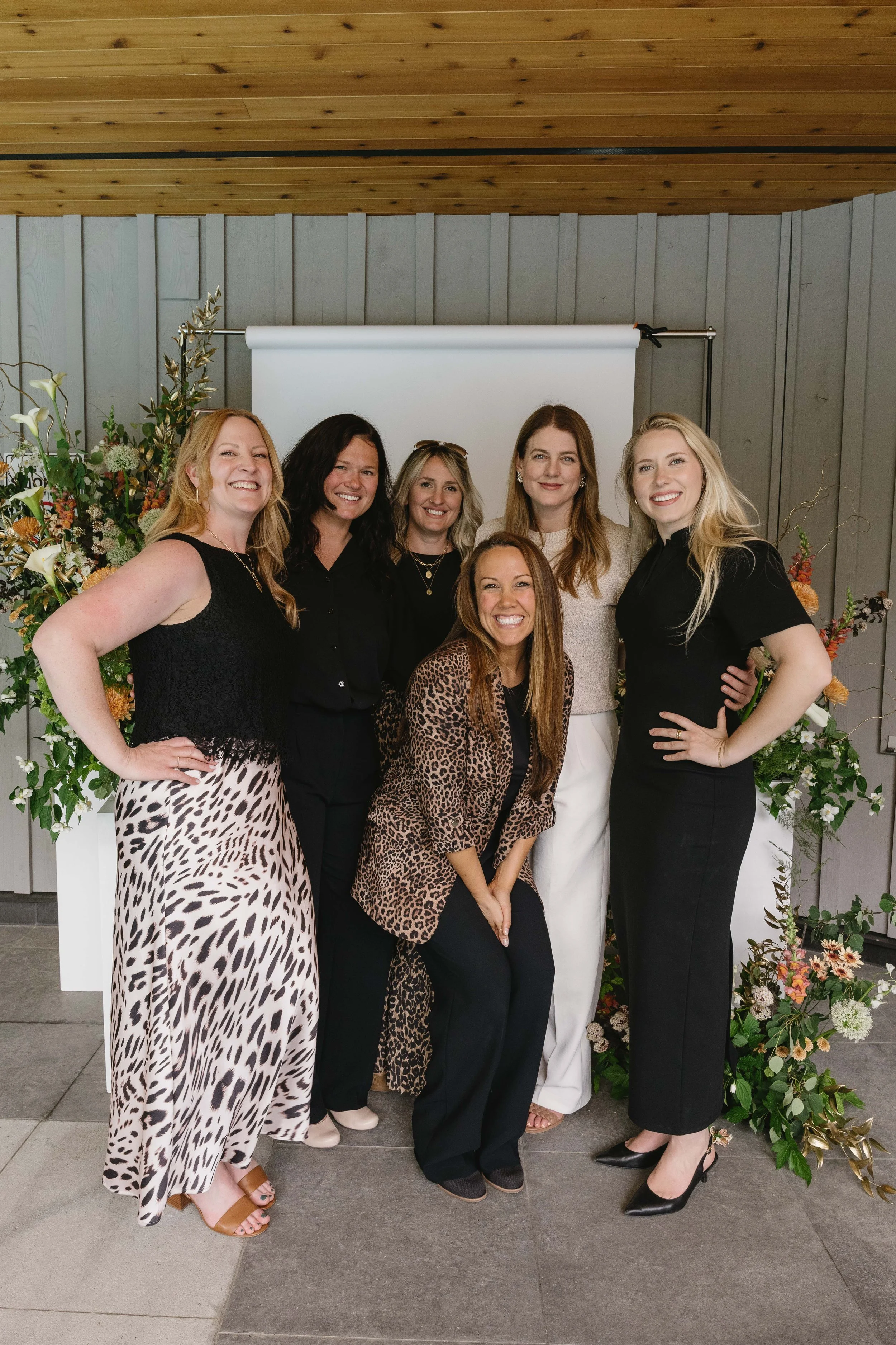 Group of seven women standing in front of a flower arrangement and white backdrop, posing for a photo indoors with a wooden ceiling and gray walls.