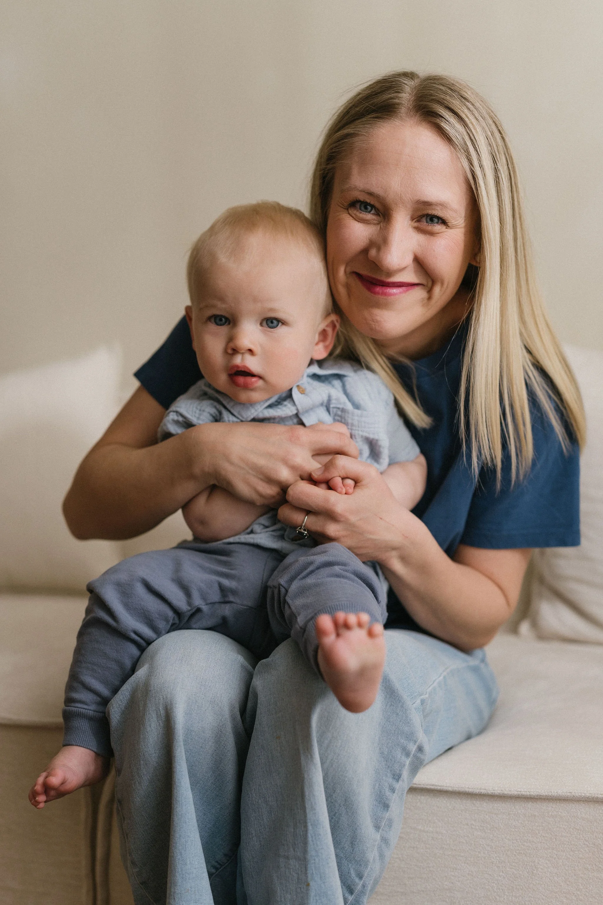 A woman with blonde hair and a blue shirt sitting on a beige couch holding a young boy with blonde hair and a light blue shirt. They are smiling and looking at the camera.