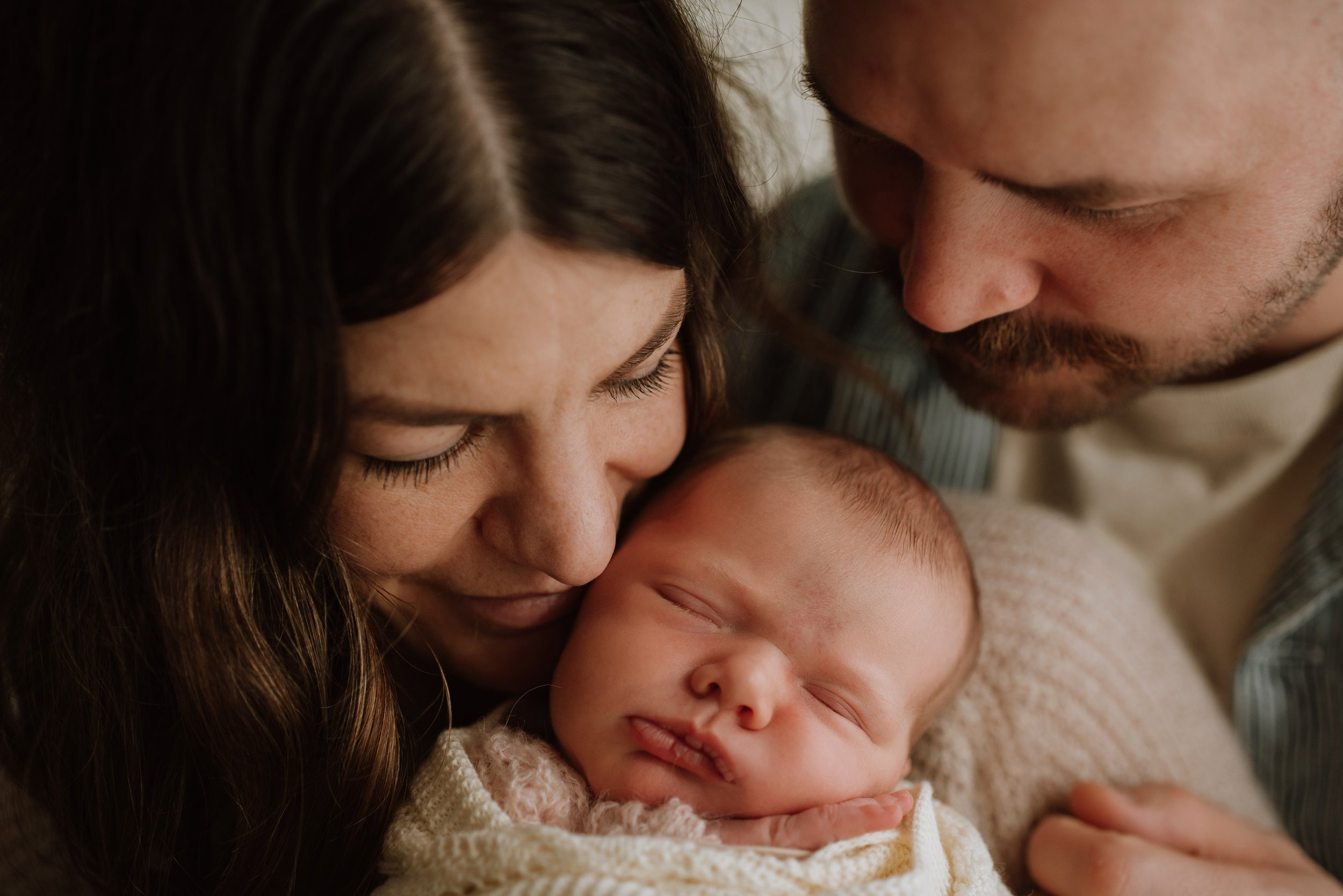 A close-up of a family, with a woman and man gently holding and kissing a sleeping baby, showing affection and tenderness.