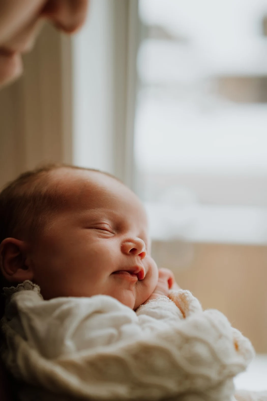 A sleeping baby with a peaceful expression, resting its head on its arms, near a window.