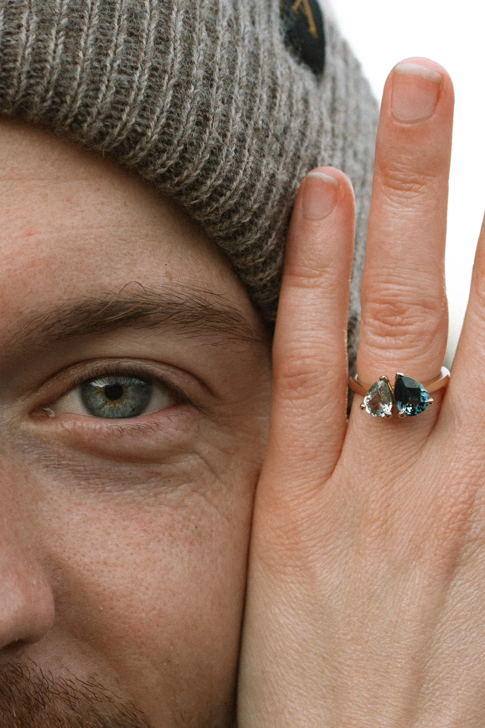 Close-up of a person's eye, part of their face, and fingers with rings, wearing a gray knit hat.