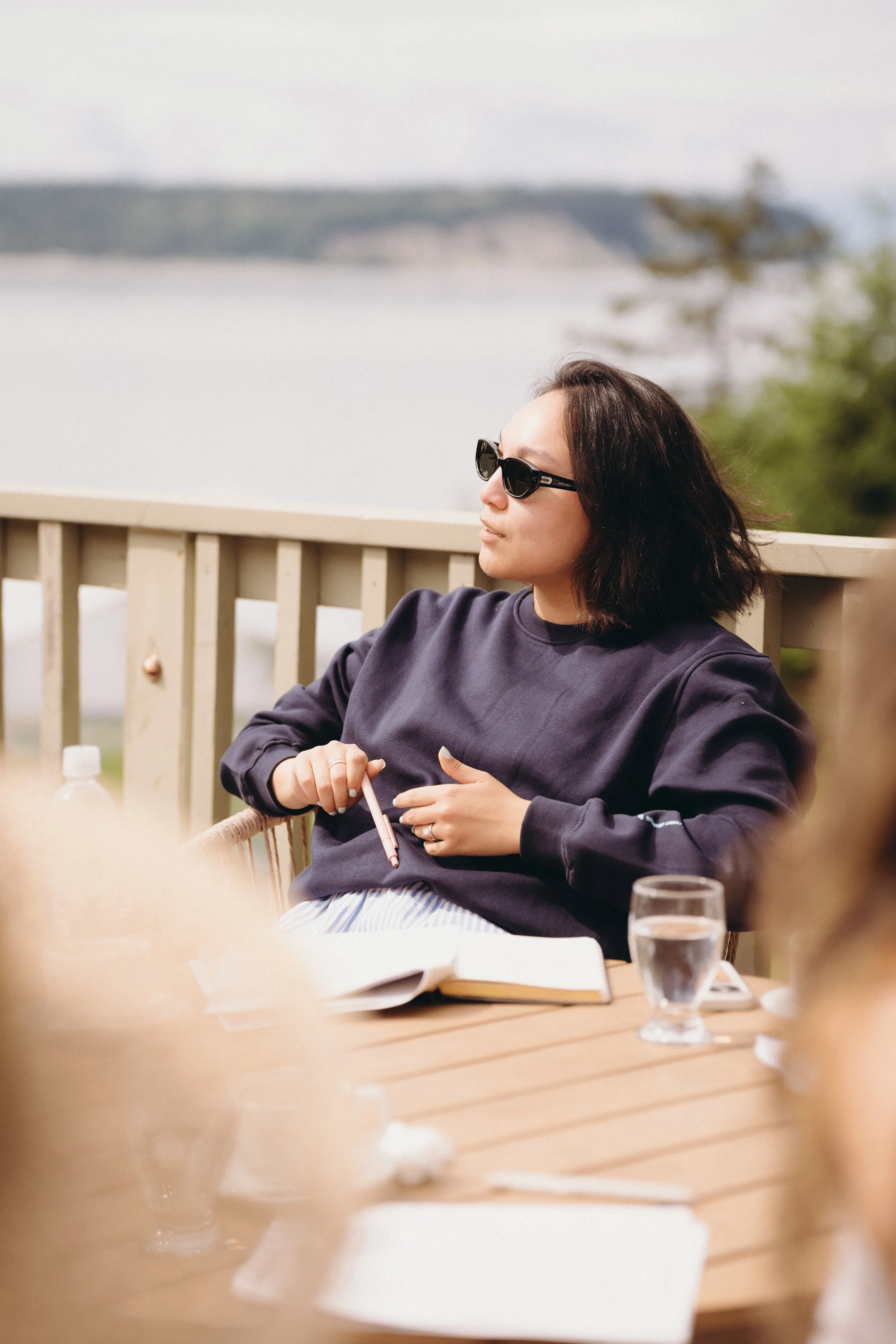 A woman with shoulder-length dark hair wearing black sunglasses and a dark sweatshirt, sitting outdoors near a body of water, with a lake and trees in the background. She is holding a pink pen and has an open notebook on the table in front of her. A 