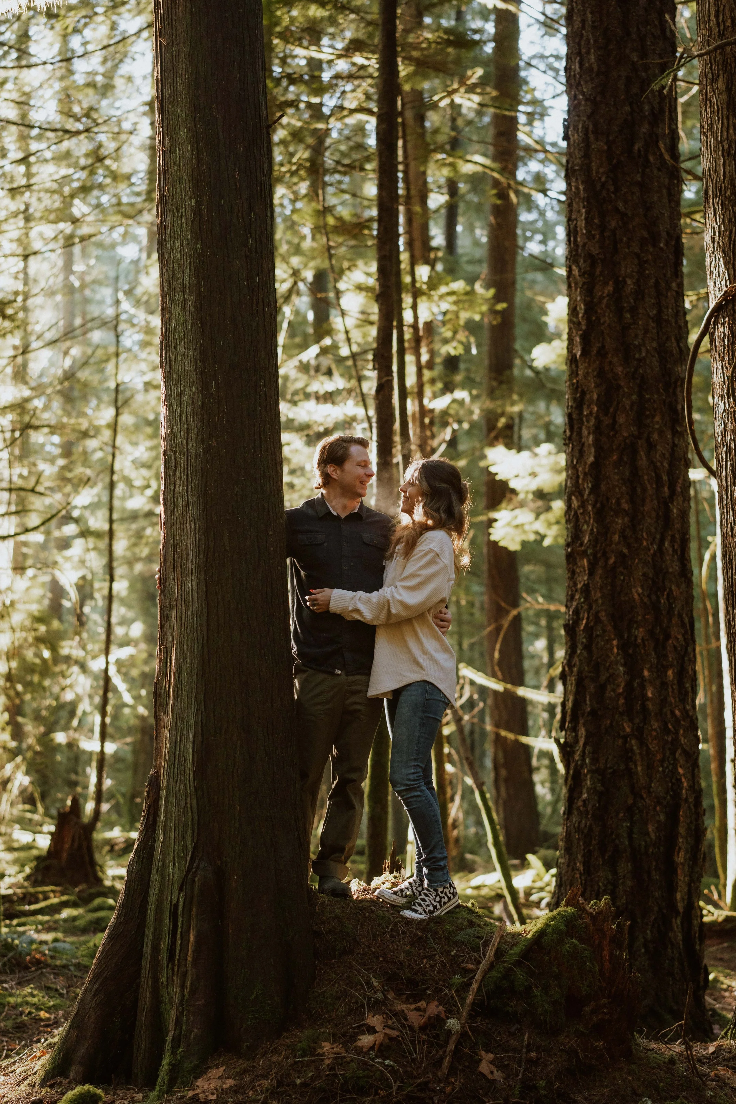 A couple standing on a mossy fallen log in a sunlit forest surrounded by tall trees, smiling and looking at each other.