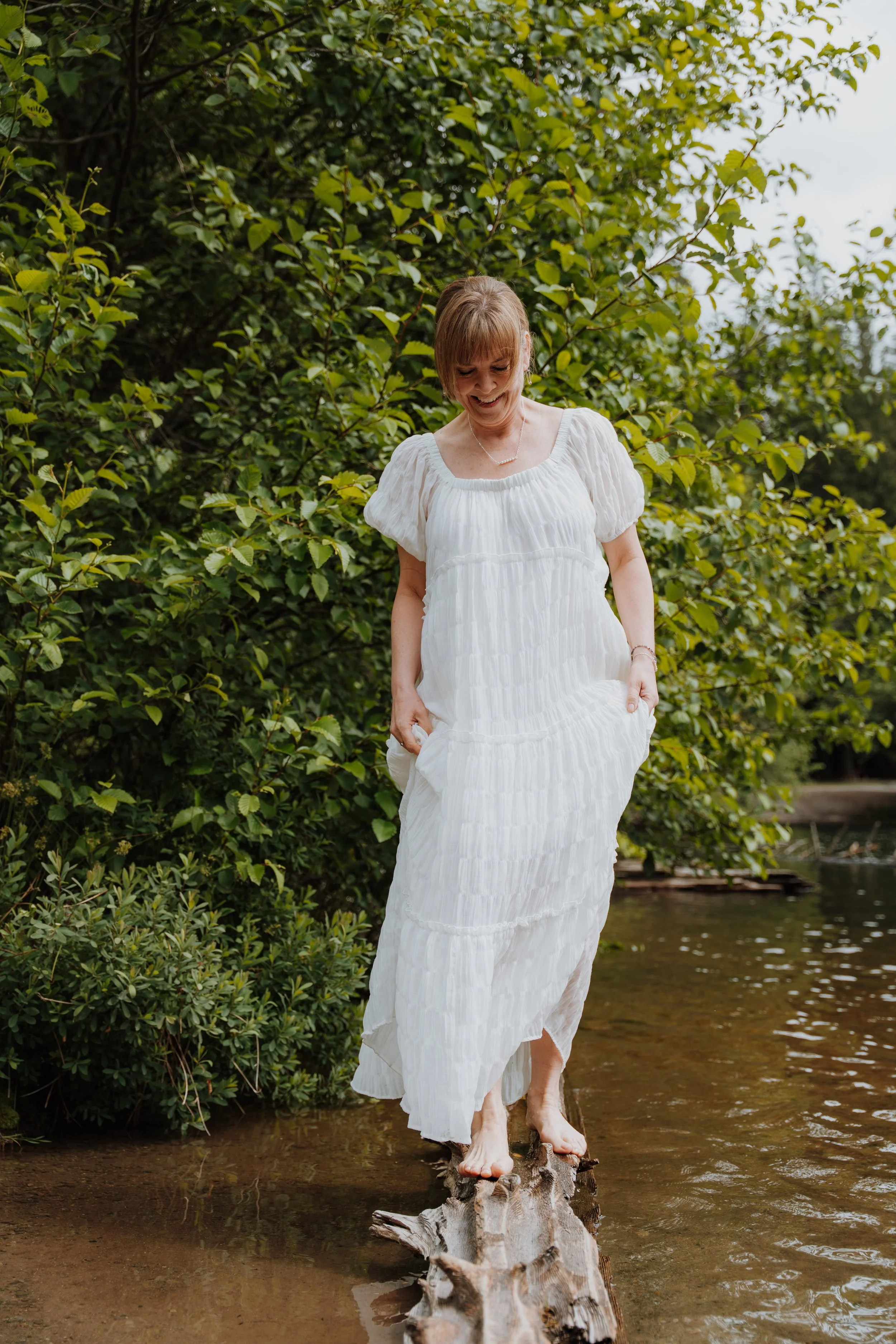 A woman in a white dress walking barefoot on a log through a shallow stream surrounded by green foliage.