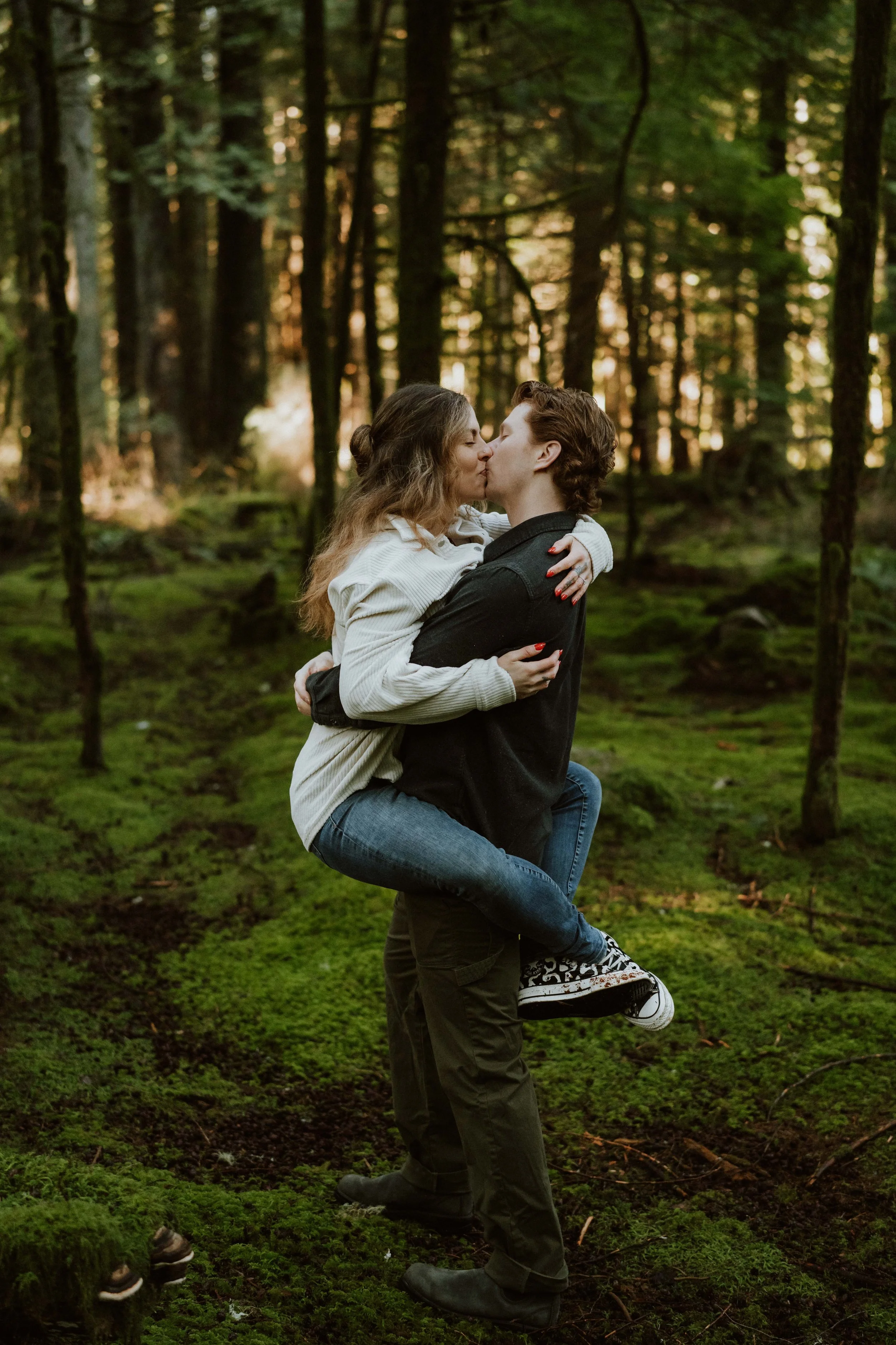A couple embracing and kissing in a forest, with the woman being carried by the man. The forest has tall trees and green moss on the ground.