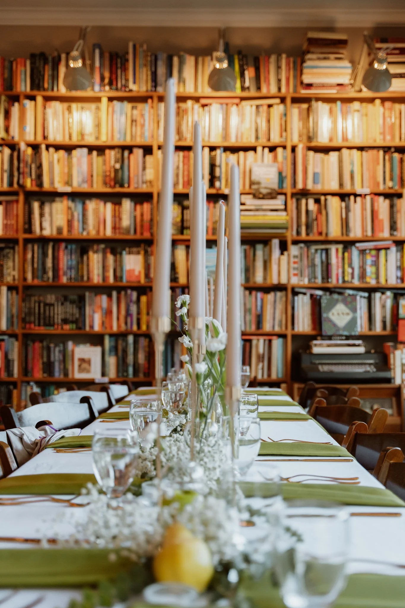 A long dining table with white tablecloth, green napkins, glasses of water, and tall white candles in the center, set in front of a large bookshelf filled with various books.