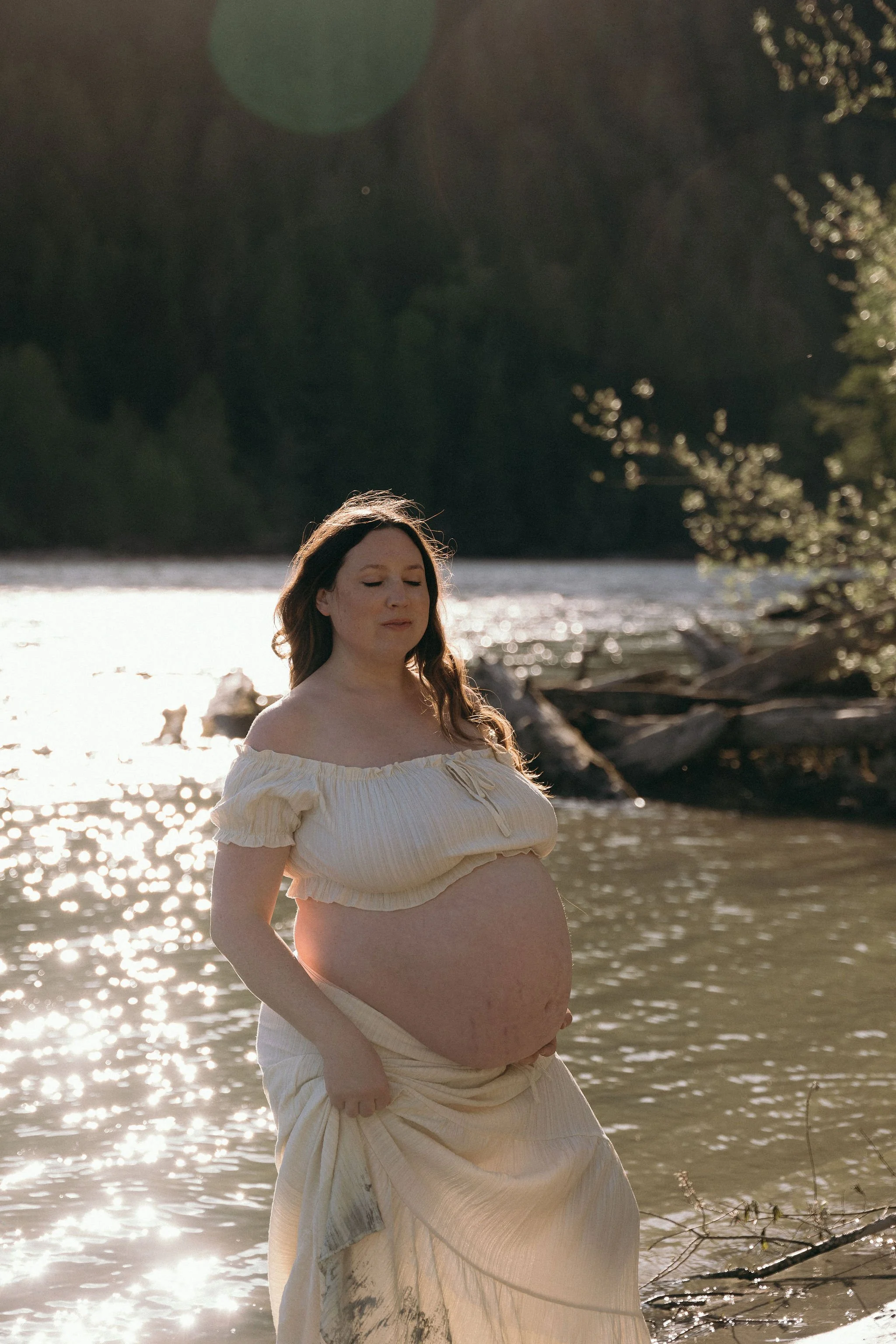 A pregnant woman with long hair stands in front of a body of water during sunset, wearing a flowing off-shoulder white dress, with trees in the background and sunlight reflecting off the water.